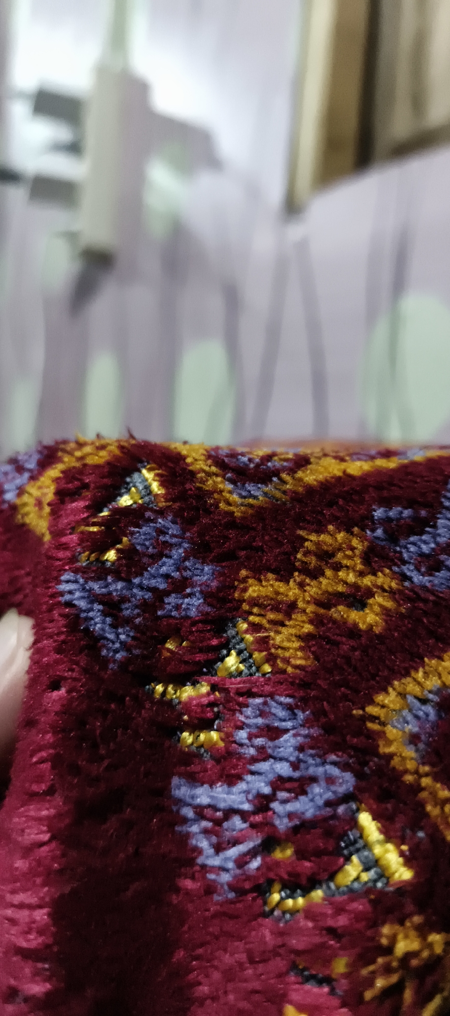 A close-up, shallow depth-of-field shot focuses on a textured, woven surface with a pattern of red, yellow, and blue threads. The material appears to be a carpet or rug, with a plush, shaggy texture. In the background, out of focus, is a blurry white object that resembles a lamp or a light fixture, situated in front of a wall with a subtle pattern of light green circles. The scene is likely indoors. The image does not provide any information about the city of Muaro Jambi, Indonesia.