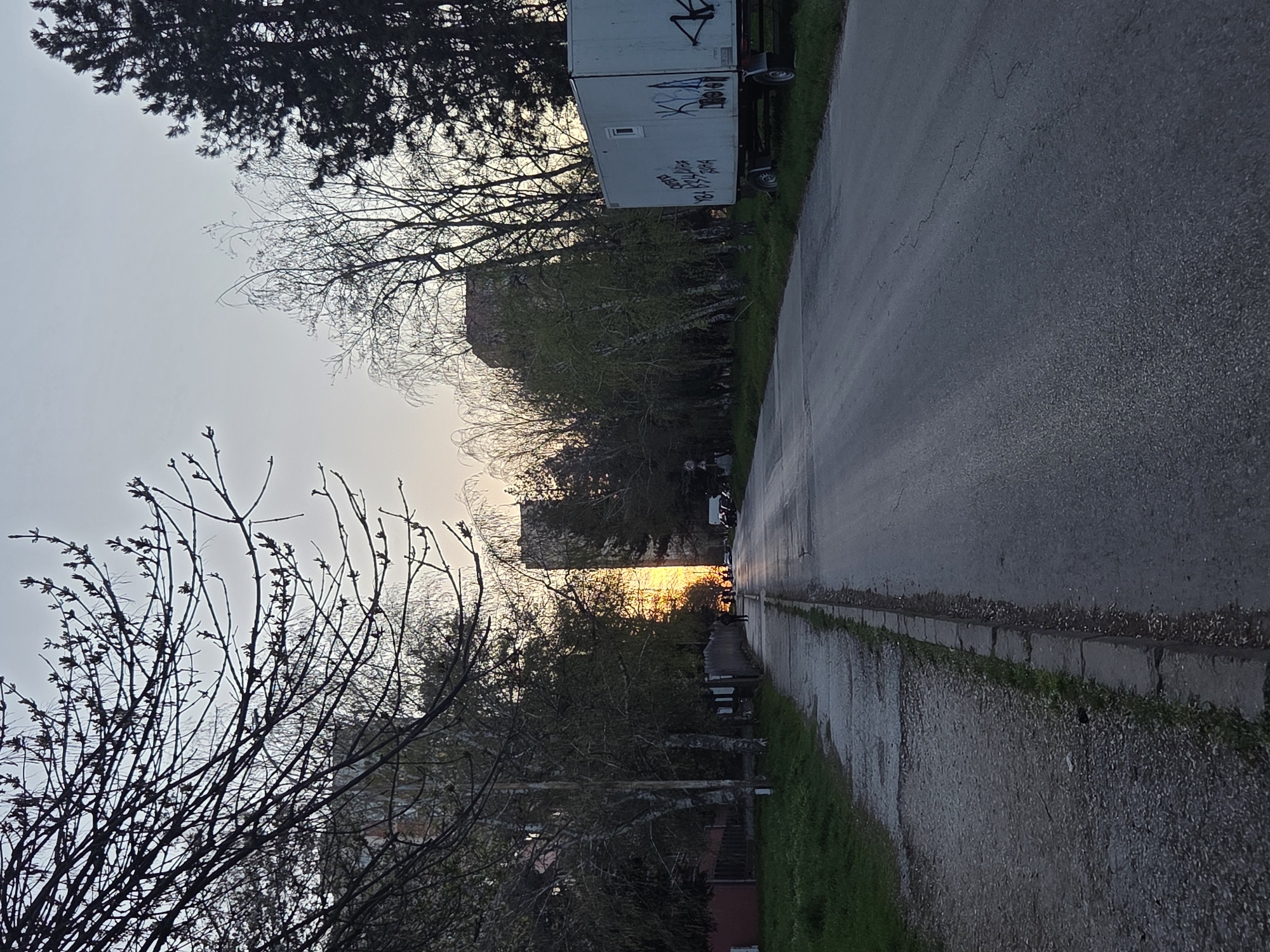 A low-angle, angled, street-level shot features an asphalt road running from the lower right to the upper center of the frame, with a concrete curb and grassy verge on the right. On the left, a bare tree with budding branches dominates the foreground. Further back, a building with a brick tower-like structure is visible, partially obscured by bare-branched trees silhouetted against the light of the setting sun. A white trailer with graffiti sits at the road's edge. The scene is likely in Pleven, Bulgaria, based on the architectural style of the brick tower. The sky is overcast, and the road surface appears wet, suggesting recent rain. People can be vaguely seen in the distance walking down the street.