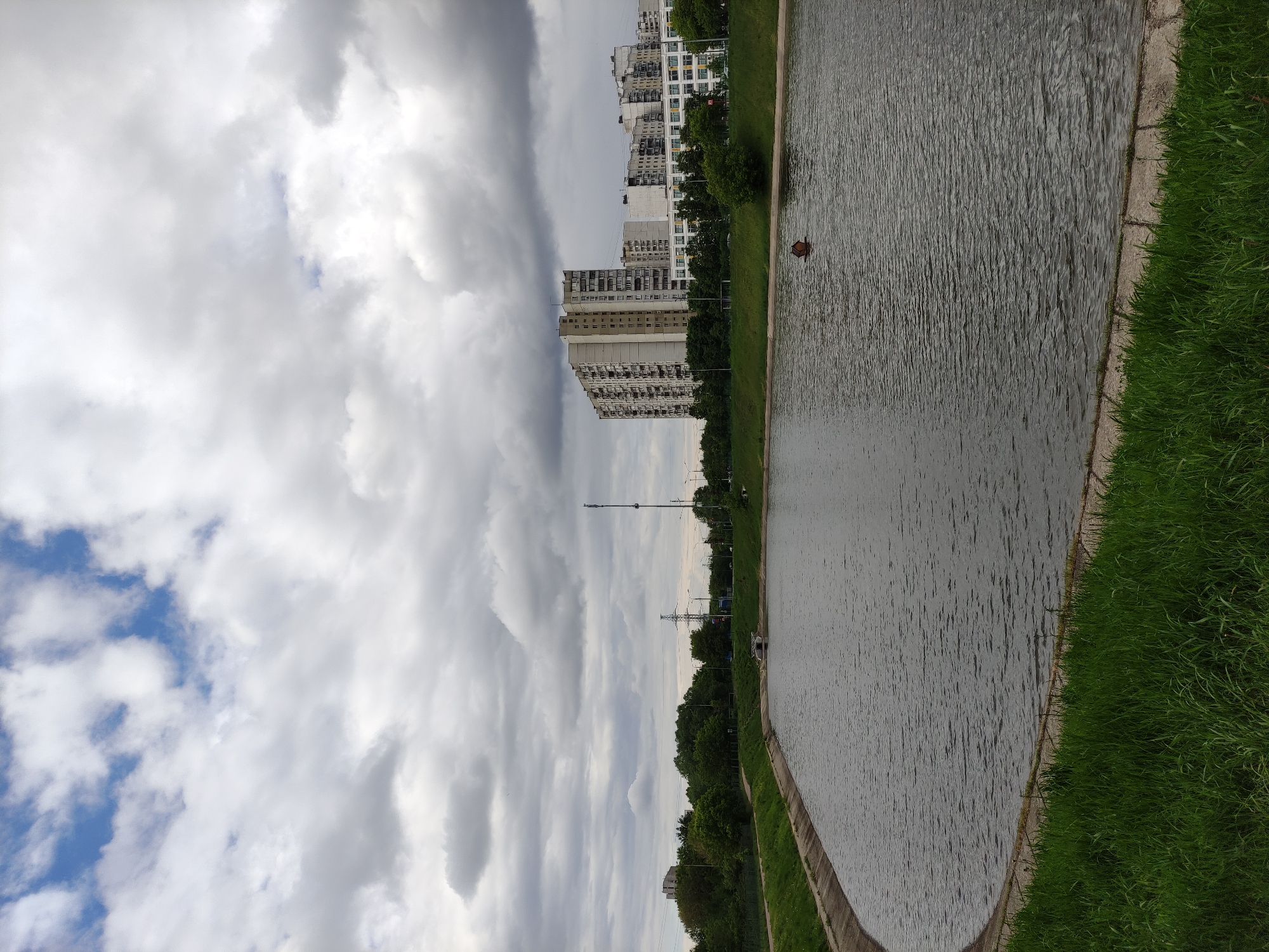The image shows a view of a canal or riverbank in a city. The water is dark, with visible ripples on the surface, bordered by a concrete edge. On the right side, a strip of green grass grows along the bank, bordering the water. In the distance, tall apartment buildings are visible, with trees interspersed along the landscape. The sky is overcast with predominantly gray clouds. A few lampposts and a communications tower are also visible in the mid-ground. There is an object floating on the water, and what appears to be a bench along the bank. The exact location within the city is indiscernible.