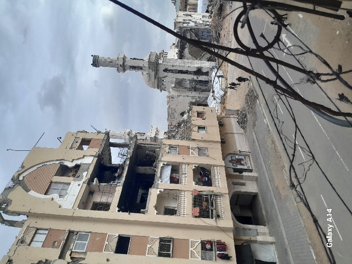 The image depicts a scene of extensive urban destruction in Khan Yunis, Palestinian Territory, under an overcast, cloudy sky, captured from an elevated perspective. In the immediate foreground, numerous dark, tangled electrical wires and a metal railing are visible on the right.
On the left side, a multi-story building, constructed of light beige material, exhibits severe structural damage. Large sections of its facade are missing, exposing the building's interior and rebar. Several floors show blackened areas, indicative of fire damage. Balconies are partially destroyed, with some railings intact, and articles of clothing are visible hanging on lower-level balconies. The upper section features a damaged ornamental arch.
Across a paved street, which contains a white directional arrow and scattered debris, stands a severely damaged mosque. Its minaret remains largely upright but shows signs of extensive damage, while the main body of the mosque is significantly collapsed into large piles of rubble and twisted metal. White canvas or tarp-like materials are discernible among the debris near the mosque.
Approximately 5-7 individuals, dressed in dark clothing, are visible walking on the street and navigating through the areas of destruction. The overall environment suggests widespread urban devastation.