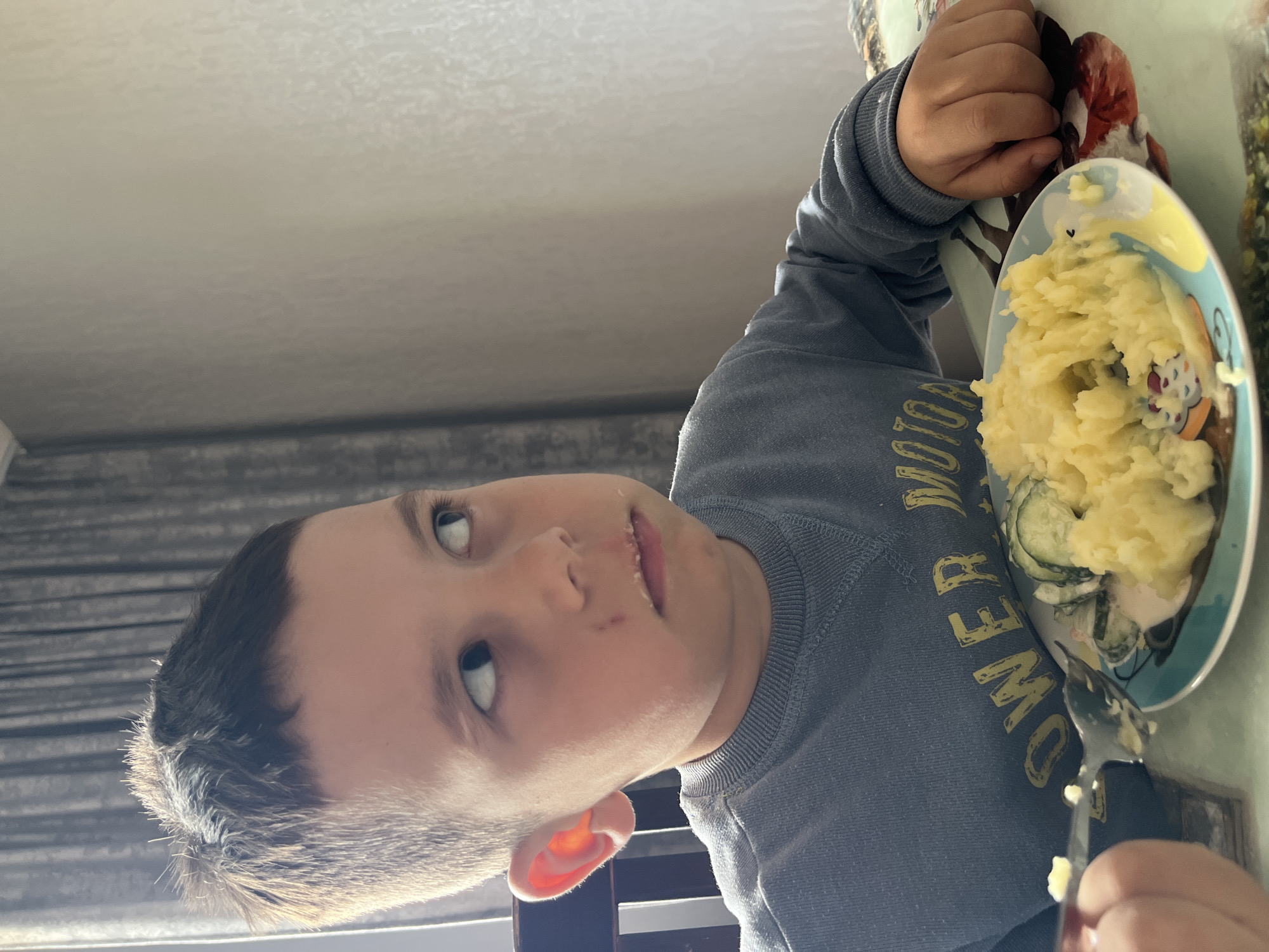 A young boy is sitting at a table, eating mashed potatoes with sliced cucumbers from a plate decorated with cartoon characters. He is wearing a gray long-sleeved shirt with yellow letters. The location appears to be indoors, possibly in a home in Chaikovsky, Russia, based on the origin of the image. The boy is looking up and slightly to the left. A spoon is in his hand, and he seems to be in the process of eating.