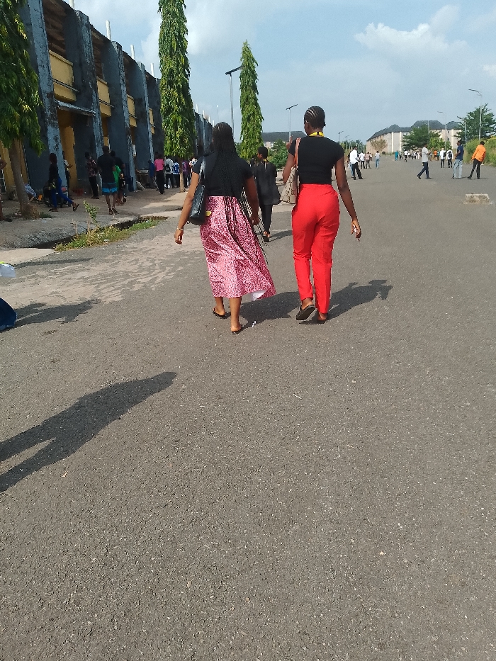 Two women walk away from the viewer on a paved road in Umueshi, Nigeria. The woman on the left wears a black top and a long pink and white skirt, while the woman on the right wears a black top and red pants. Both carry handbags. Numerous people are visible in the distance. A large building with vertical architectural features is on the left, and tall, slender trees line the road. The day is sunny, with long shadows cast on the asphalt.