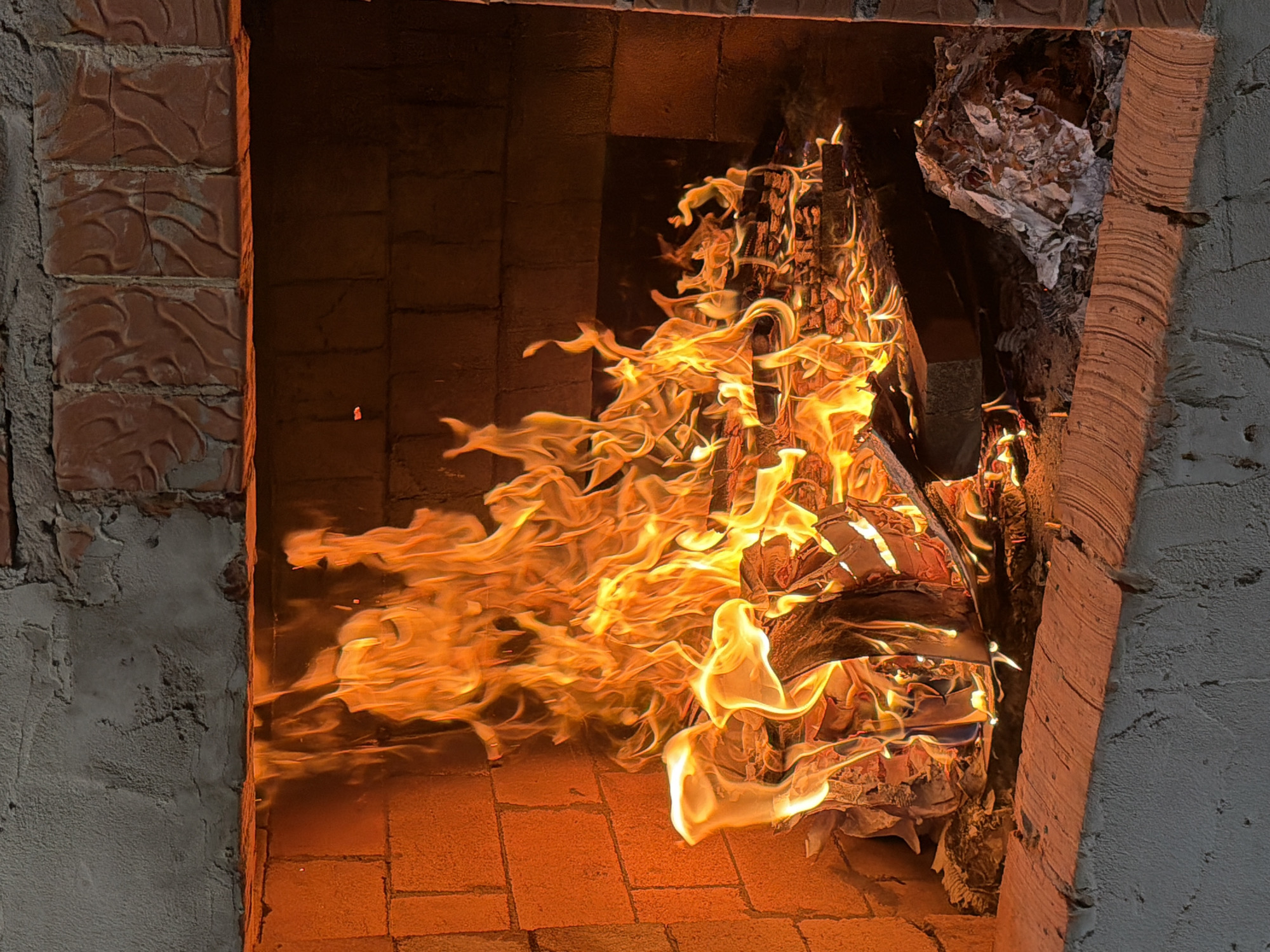 The image depicts a fire burning inside a brick fireplace. The flames are large and orange, reaching towards the top of the fireplace. The fire is consuming wood and paper materials. The interior of the fireplace is constructed of brick, while the exterior appears to be a combination of brick and mortar. The exact location of the fireplace in Chaikovsky, Russia, cannot be determined based on this single image.