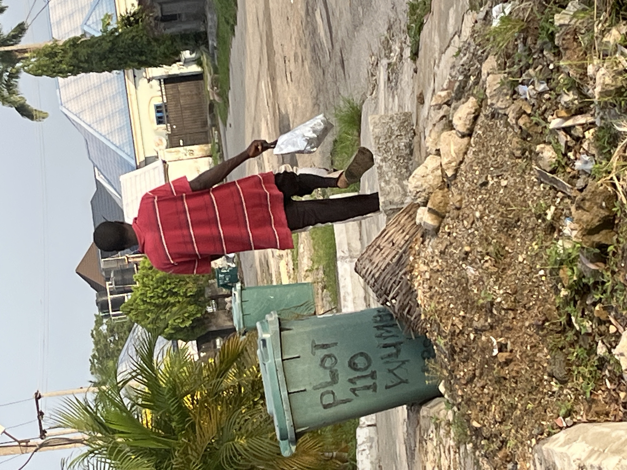 The image depicts an outdoor scene, likely in a residential area of Abuja, Nigeria, during the day. In the foreground, a green trash bin is visible with markings that appear to be "PLOT 110" and "WYUMB." Adjacent to the bin is a pile of dirt and debris, including rocks and scattered white objects that might be pieces of paper or plastic. A man, wearing a red and white striped t-shirt and dark pants with a white stripe, walks away from the camera towards the right. He is carrying a silver-colored bag in his right hand. In the background, there are houses with light-colored walls and blue-tiled roofs, some with visible water tanks on top. Lush green vegetation, including palm trees and other bushes, are scattered throughout the scene, suggesting a tropical or sub-tropical environment. The ground appears to be paved in some areas, with a dirt road or path visible further back. The overall impression is of a slightly unkempt urban or suburban neighborhood.