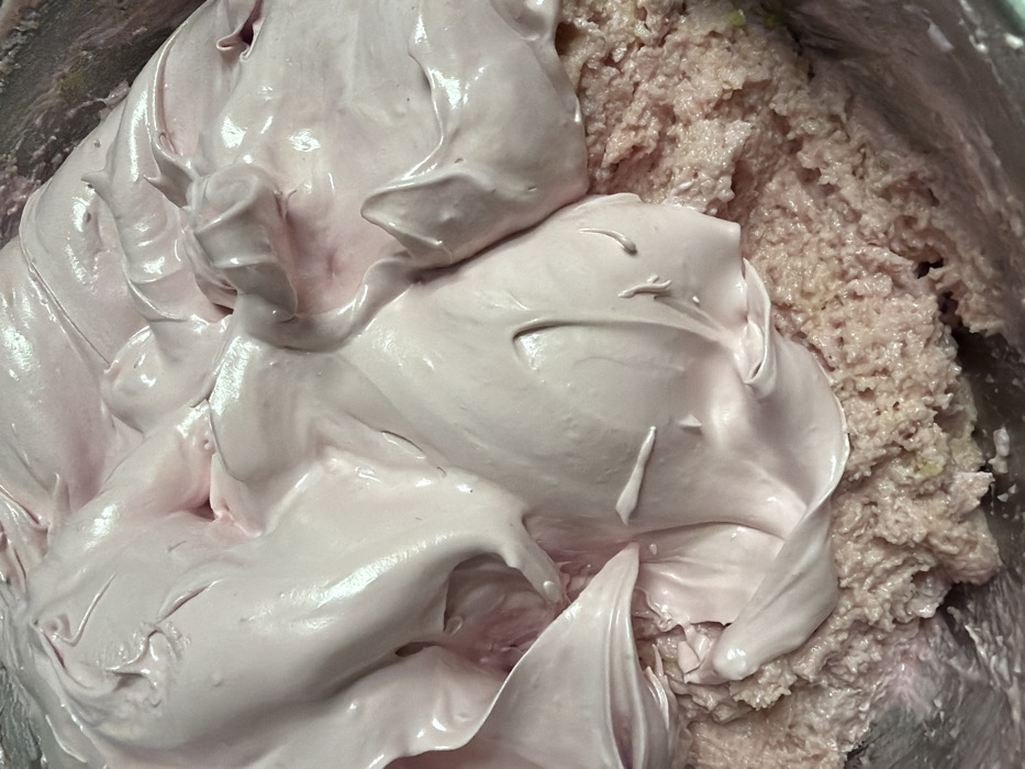 A close-up reveals a mixing bowl containing two distinct textures. One side features a mass of smooth, light pink frosting with swirling peaks. The other side consists of a coarsely textured, pale pink mixture speckled with lighter-colored flecks. The frosting and mixture are partially blended at the interface, suggesting they are being incorporated together. The overall impression is one of a dessert or baking preparation in progress.