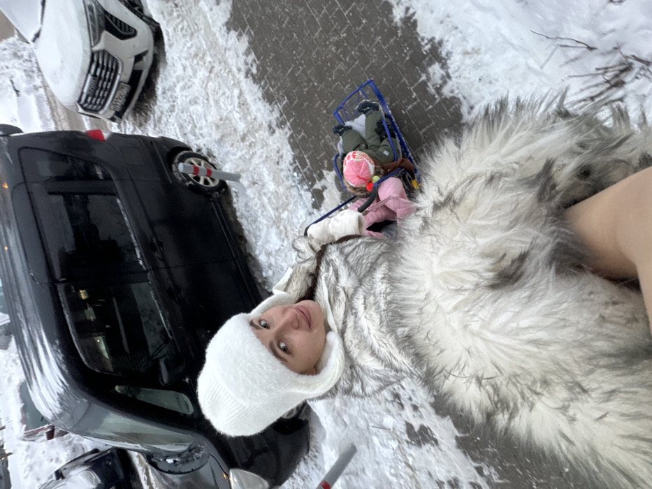 The image presents an outdoor winter scene set in Odintsovo, Moscow Region, Russia. In the foreground, an adult female with light brown hair and a smiling expression is oriented towards the camera. She is attired in a white knitted winter hat and a thick, light gray and white faux fur coat. Her right arm, gloved in white, extends rightward, suggesting engagement with an unseen object. To her right, two small children are situated in a blue metal-framed sleigh. The child on the left wears a pink winter suit and hat, while the child on the right wears an olive green winter suit and a pink and white striped hat with a pom-pom. The ground consists of a partially snow-covered gray paver walkway alongside a snow-laden vehicle area. A dark-colored minivan/van is parked on the left, and a white Haval SUV bearing Russian license plate P110OC 190 is parked further right. Red and white striped flexible bollards delineate the vehicle area from the walkway. The scene is illuminated by daylight under snowy conditions.