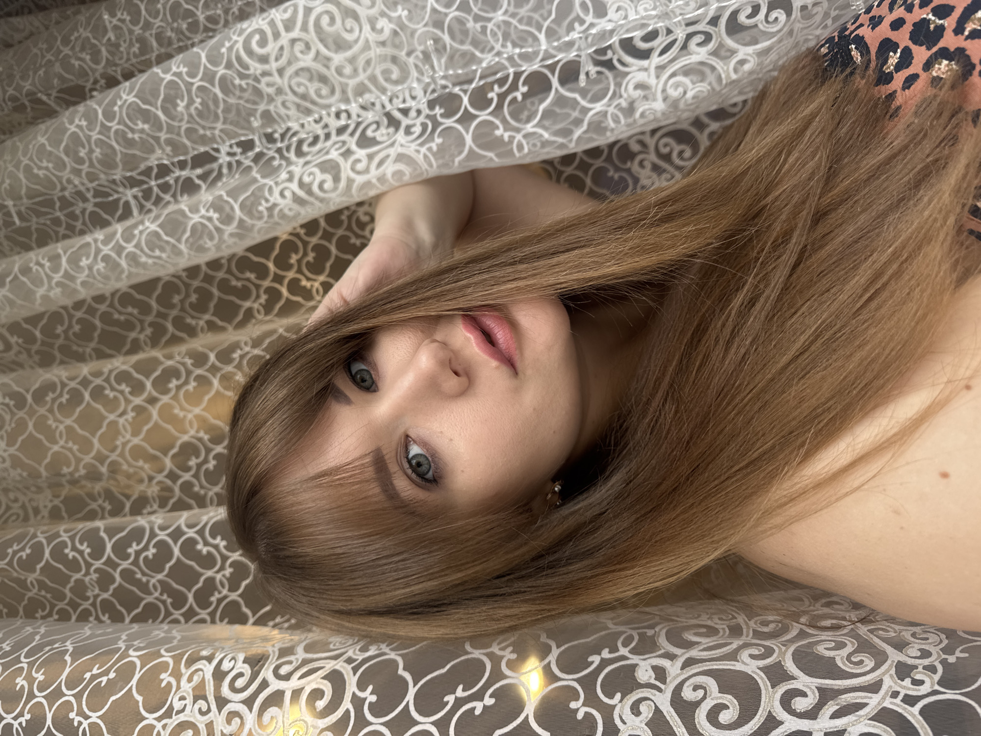 A medium shot shows a young woman with long, brown hair and bangs, positioned near a window with sheer, patterned curtains. She is looking directly at the camera with a neutral expression. Her right hand is resting on the back of her head, partially obscuring her hair. She is wearing a leopard-print top. The background consists of two layers of curtains, one behind the other. The lighting suggests an indoor environment, possibly a home. The image does not contain enough information to determine a specific location in Chaikovsky, Russia, or any forensic details beyond what is visible.