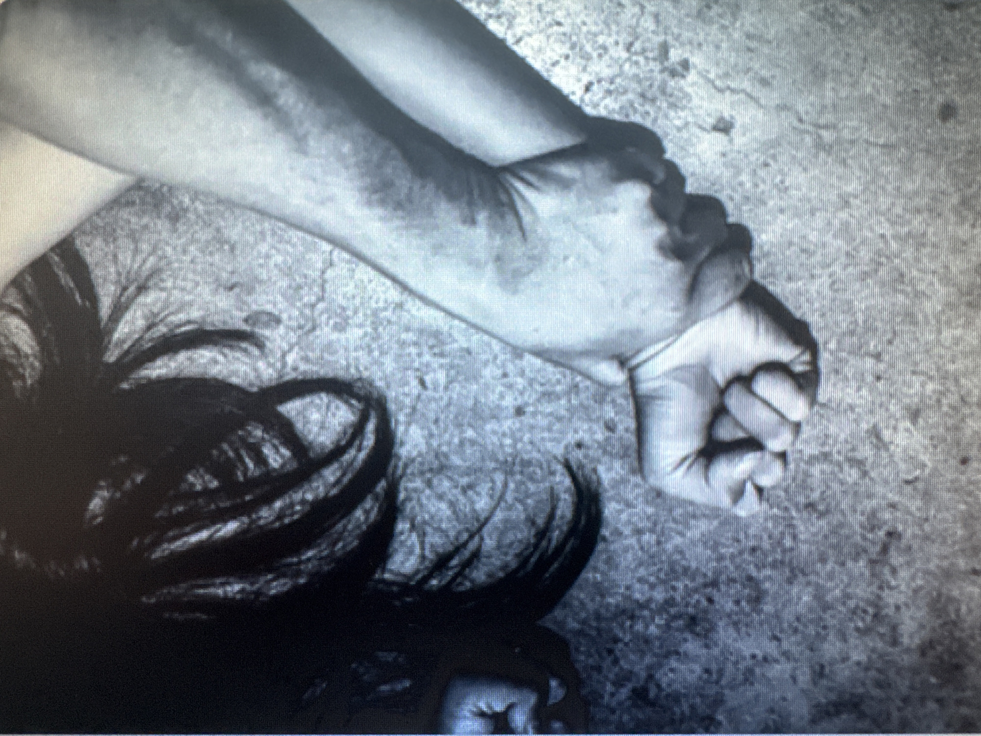 The image displays a close-up, black and white shot of two arms. One arm, appearing to be an adult's, is firmly gripping the wrist of another arm. The gripped wrist is clenched into a fist. The background is a rough, textured surface, possibly concrete. The lighting is dim, creating shadows and highlighting the textures of the skin and the ground. The top right corner shows a portion of dark hair, suggesting a person is lying down. No specific location within Protasovo, Russia can be identified from the visual information provided.