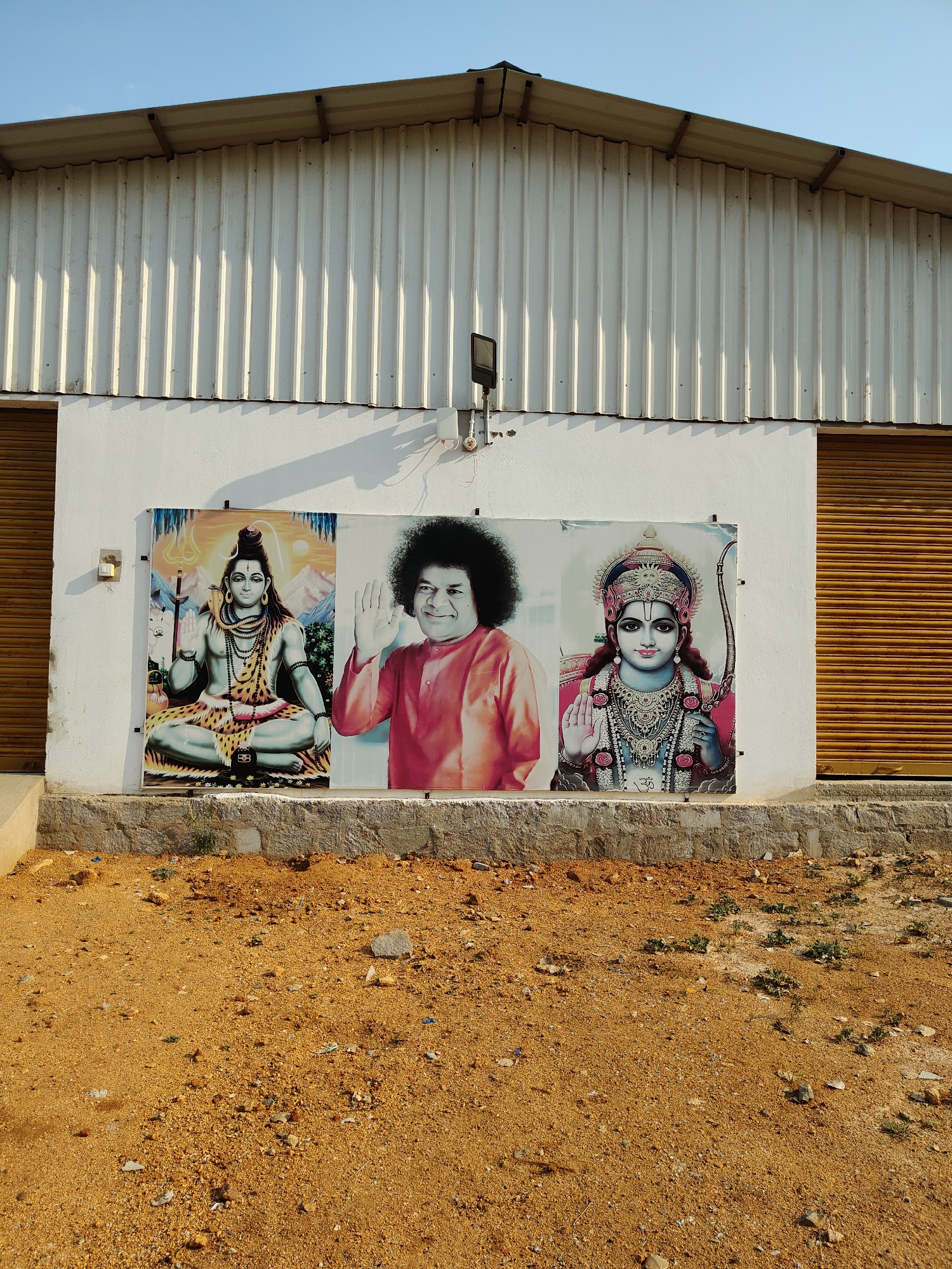 The image shows a building exterior in Pedda Kammavaripalli, India. The building has a white, corrugated metal roof and a white wall. Affixed to the wall are three adjacent posters depicting religious figures: Shiva, Sai Baba, and Rama. Below the posters is a low retaining wall. The ground in front of the building is dirt and gravel. The sky is clear and blue. Two roll-up doors flank the posters. A light fixture is mounted on the wall above the posters.