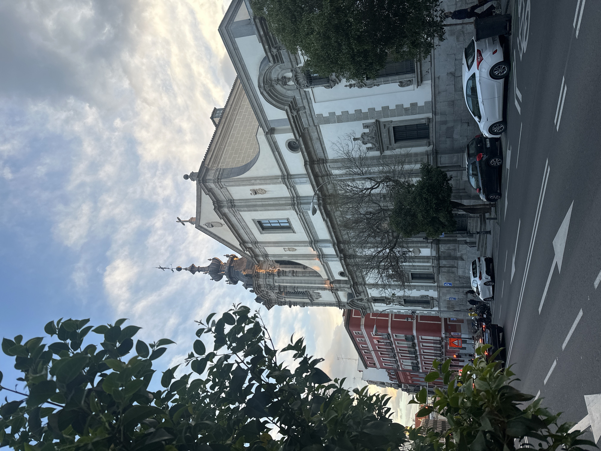 The image depicts a street scene in Madrid, Spain, featuring the Royal Basilica of San Francisco el Grande. The basilica is a large, white and gray stone building with baroque architectural elements. A tall, ornate steeple rises from the church.
The street runs parallel to the basilica. There are several cars parked along the side of the road, including a white hatchback and a black sedan. There are also several motorcycles parked on the street. A red and white building stands adjacent to the church.
In the foreground, lush green foliage from trees partially obscures the lower portion of the image. The sky above is partly cloudy with patches of blue visible. The light suggests it may be late afternoon or early evening. A person is visible standing near the white vehicle, interacting with the vehicle. Road markings indicate traffic flow.