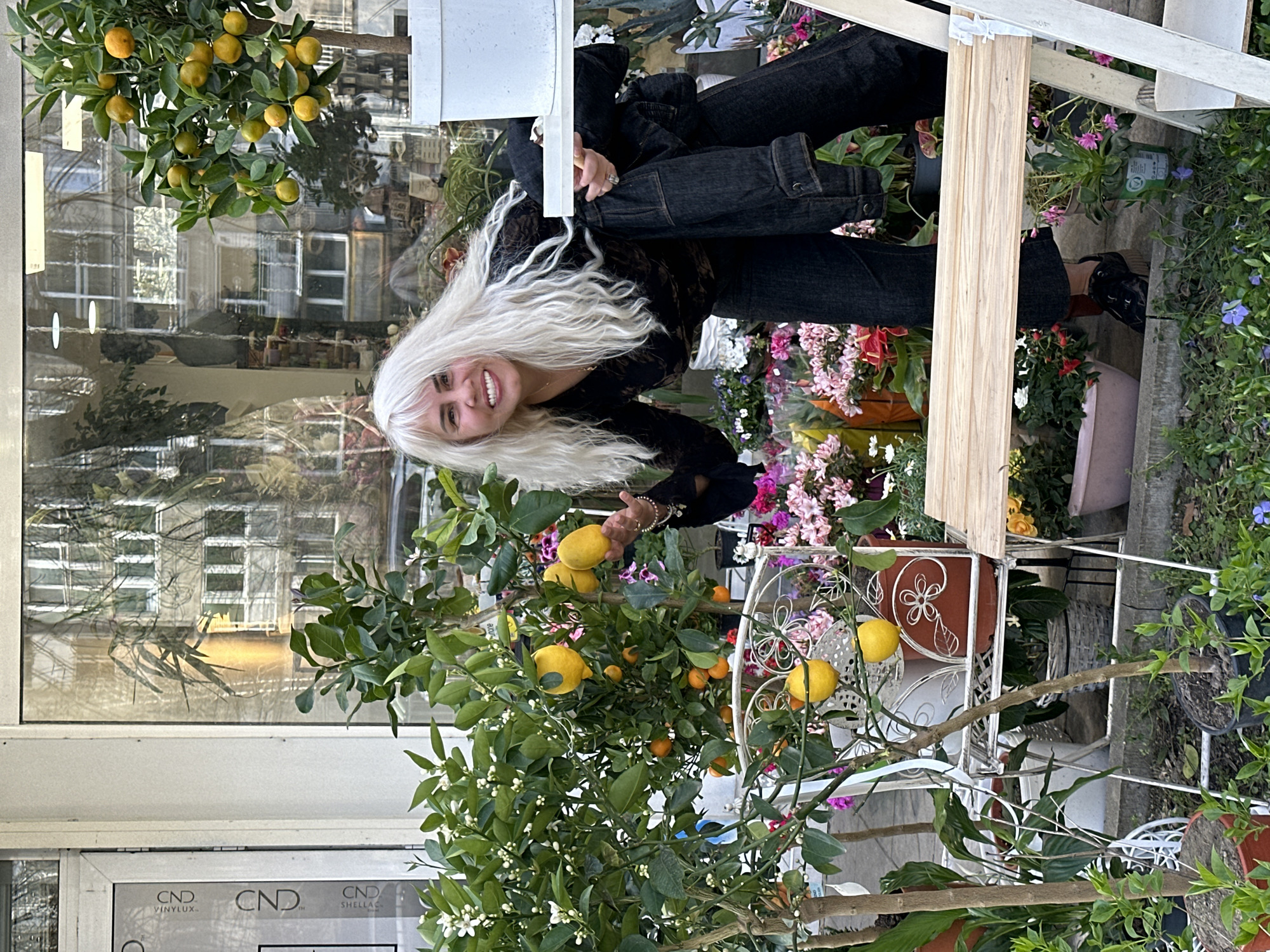 A vertical shot reveals a smiling woman with platinum blonde hair positioned near a citrus tree, presumably in Sofia, Bulgaria. The woman is wearing black clothing and standing beside a citrus tree laden with yellow lemons and small oranges. The tree is in a white, ornate metal stand among other plants. Behind the woman and the tree, a building with large windows is visible, reflecting the surrounding urban environment. The "CND" brand is visible on the building. Additional potted plants with colorful flowers contribute to a lively atmosphere. A portion of a wooden structure is also visible in the frame.