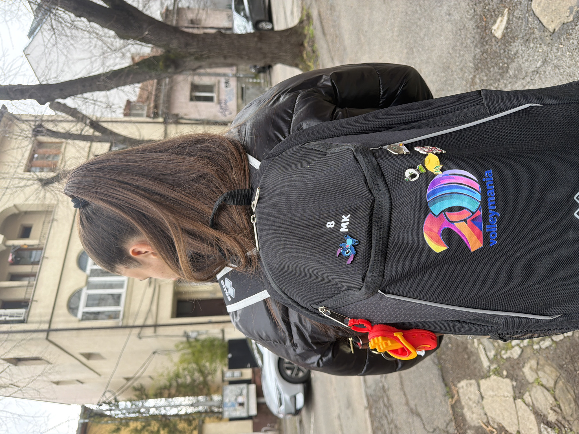 The image captures a person wearing a black puffy jacket and a black backpack with pins and a "volleymania" logo, standing on a street in Sofia, Bulgaria. The person has brown hair pulled back in a ponytail and is looking down. The backpack is adorned with several pins, including a cartoon character and a lemon. A small pouch on the backpack has the initials "MK" and the number "8." Attached to the backpack's zipper is a red and yellow plastic toy.
The background includes a tree with bare branches, buildings with windows, and a car parked on the side of the street. The environment appears to be an urban area with typical European architecture. The overall impression is a casual, everyday scene on a cloudy day in Sofia.