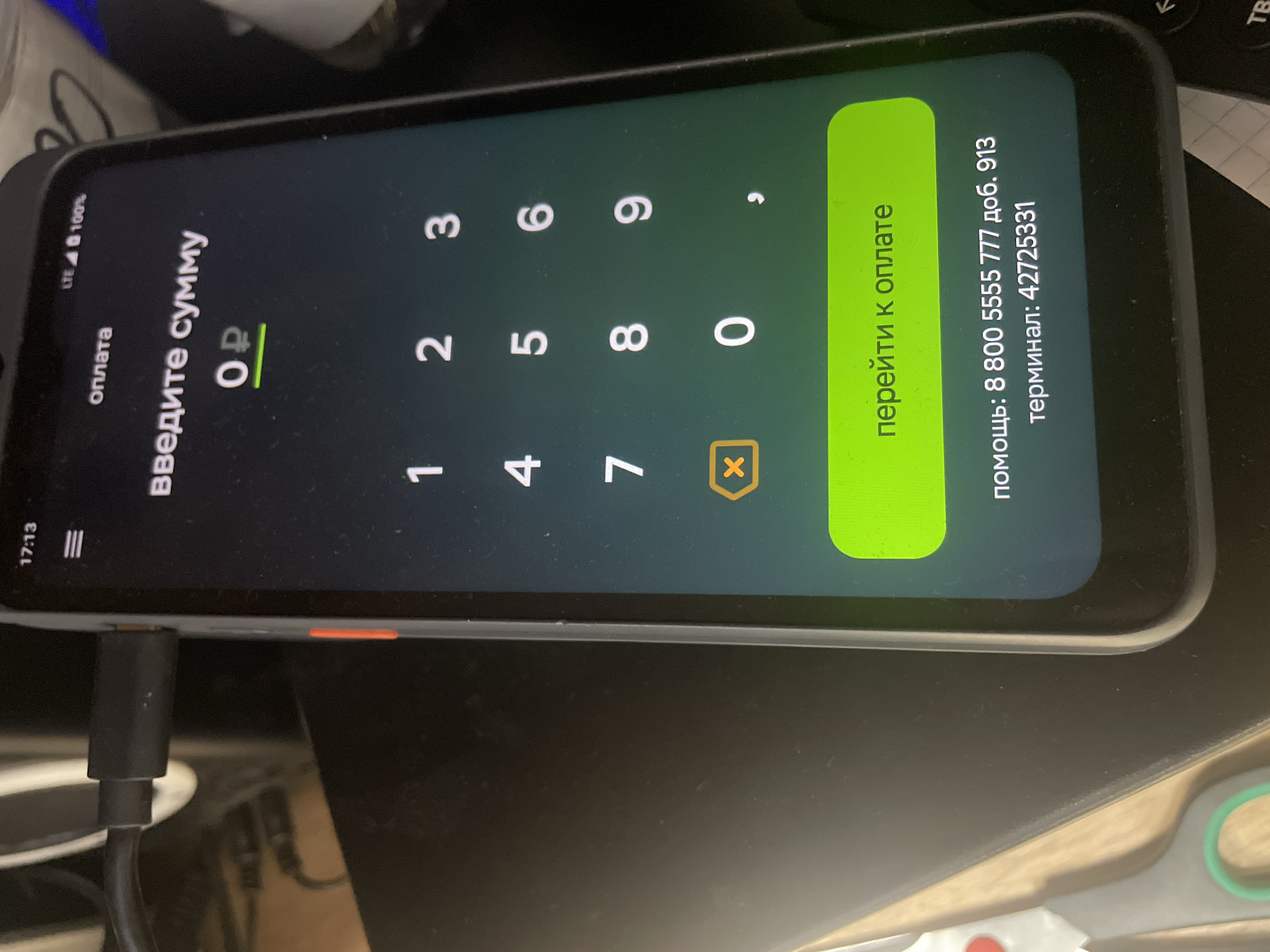 The image shows a close-up, rotated view of a smartphone screen displaying a payment interface. The screen is dark with green and white text and graphics. The primary subject is the smartphone, which is plugged into a black cable at the bottom. The phone is displaying a numeric keypad and input fields.
The interface text is in Russian. At the top left, "LTE 100%" indicates the signal strength and battery level. Below that, "оплата" (payment) is visible, followed by "введите сумму" (enter amount). The amount field shows "0 ₽", indicating zero rubles, with a horizontal line beneath it for input. The numeric keypad has digits 1 through 9 arranged in a standard layout, with 0 to the right of 7, 8, and 9. A comma key is to the right of 9, and an 'X' symbol within a yellow box is below 7, 8, and 9.
To the right of the keypad, a large, bright green button displays the text "перейти к оплате" (proceed to payment). Further to the right, there is information about assistance and a terminal number: "помощь: 8 800 5555 777 доб. 913" (help: 8 800 5555 777 ext. 913) and "терминал: 42725331" (terminal: 42725331).
The scene appears to be indoors, with the phone placed on a dark surface, possibly a desk or table. There are hints of other objects in the background, out of focus, including what looks like part of a keyboard and some lighter colored materials. The time displayed on the phone's status bar is "17:13". There are no people visible in the image. The overall impression is a functional, straightforward display of a mobile payment application.