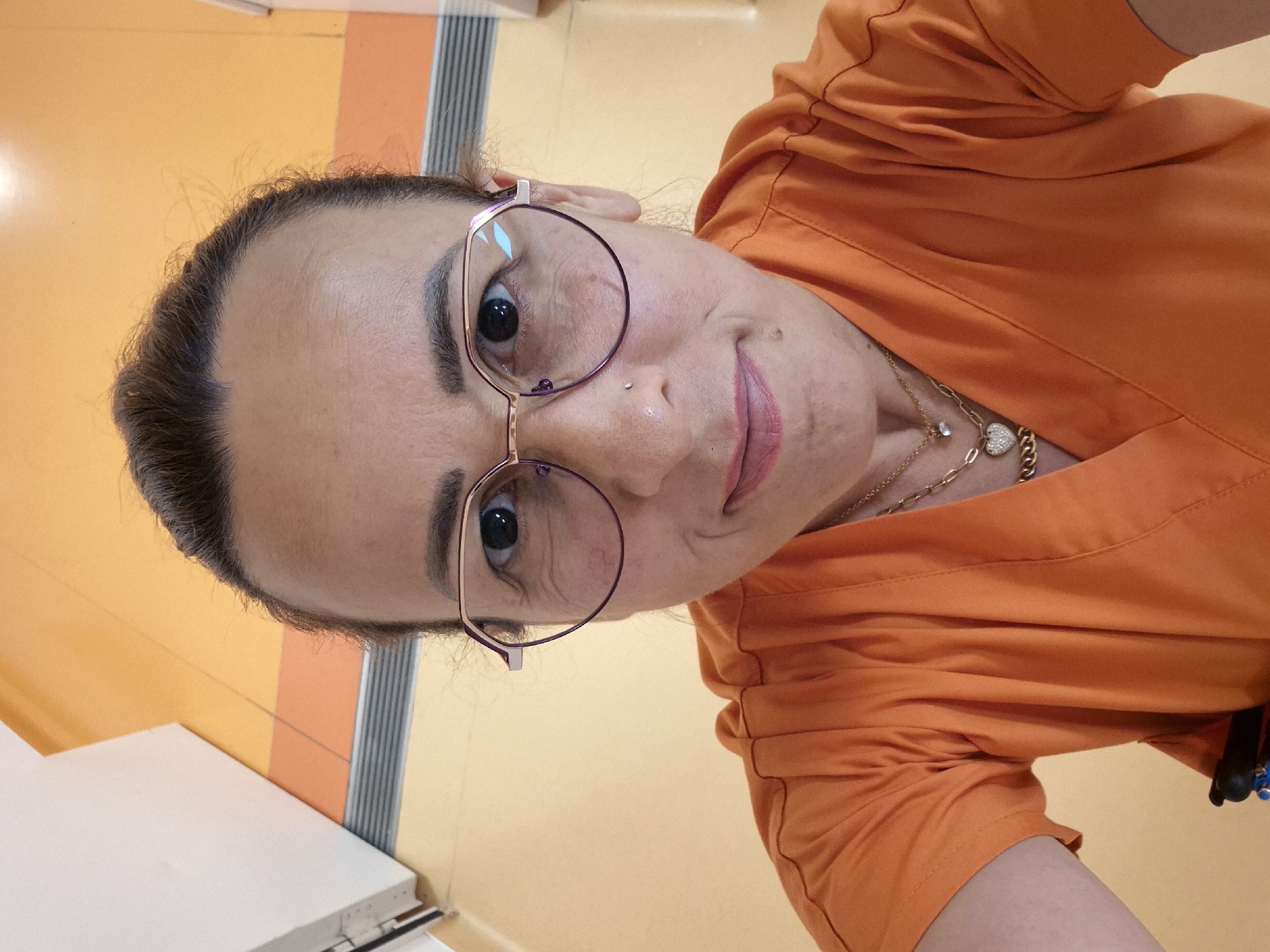 A medium shot captured from a slightly high angle shows a woman with her hair pulled back and wearing glasses and an orange v-neck scrub top. The woman is looking directly into the camera, and her head is tilted slightly to her left. She is wearing a gold chain with two pendants: one a small, clear stone, and the other a heart-shaped charm encrusted with small diamonds. The background is a bright orange, and there is a white wall on the left side of the frame. The environment appears to be an indoor setting, possibly a medical or clinical facility, given the presence of the scrub top and the clean, institutional-looking walls and flooring.