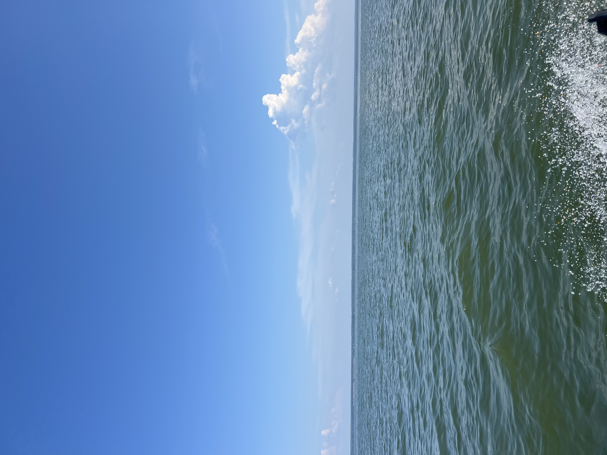 An outdoor, eye-level, medium shot captures a serene scene of a vast body of water under a bright blue sky. The water, with its gentle ripples and a hint of green hue, stretches towards a distant, flat horizon. In the upper right quadrant, a dramatic, voluminous white cloud stands out against the clear sky, while thinner wisps of clouds are scattered across the expanse. The foreground on the right side of the frame shows a frothy spray of water, suggesting movement, possibly from a boat. The location is identified as the city of Borovoe, Russia. No people are visible in the image.