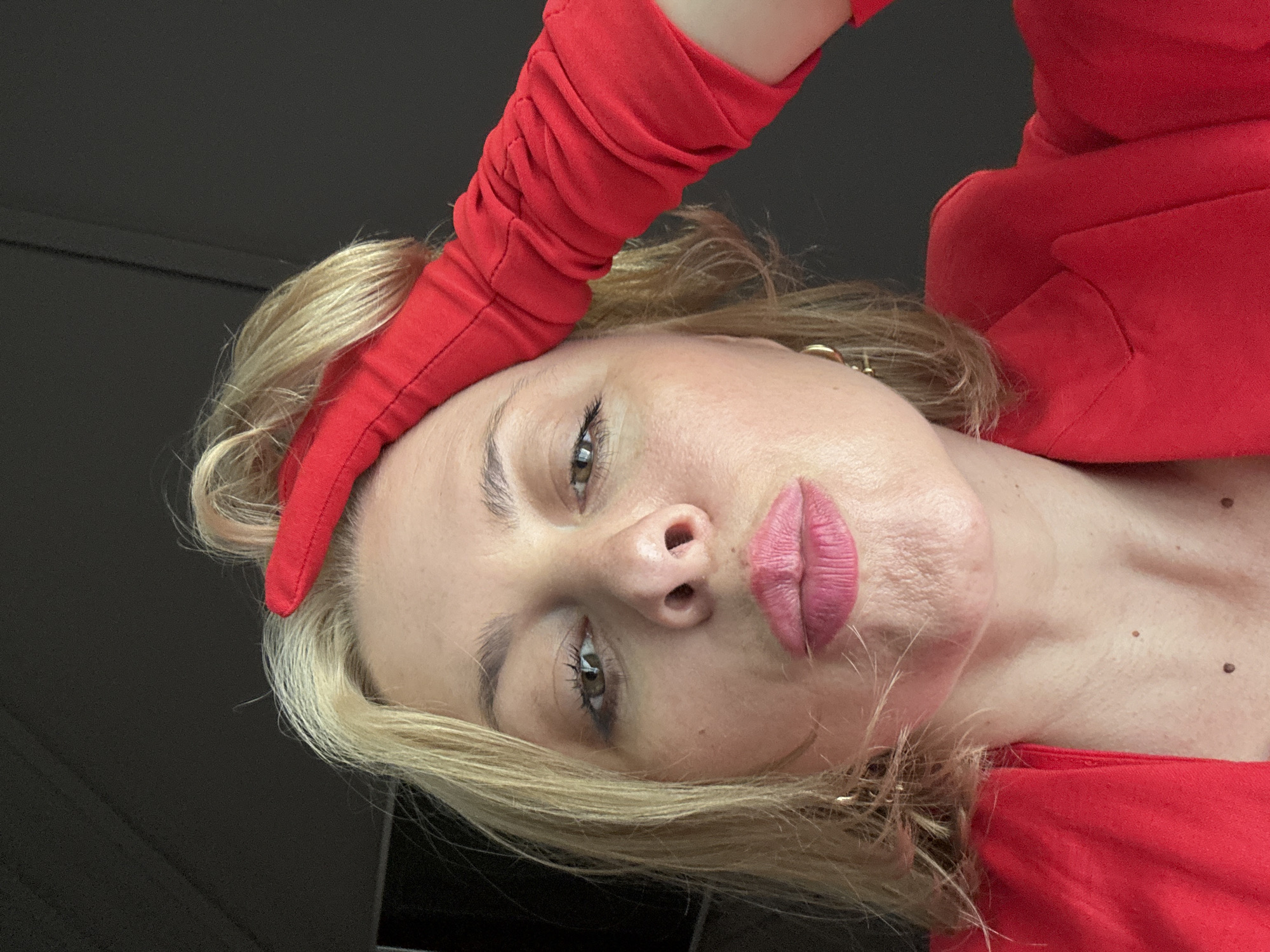 A woman with blonde hair and pink lipstick is looking at the camera and holding her forehead with a red glove. She is wearing a red jacket. The background is dark. The photo appears to be taken indoors. There is no information or visual evidence in the image to suggest it is located in Sochi, Russia.