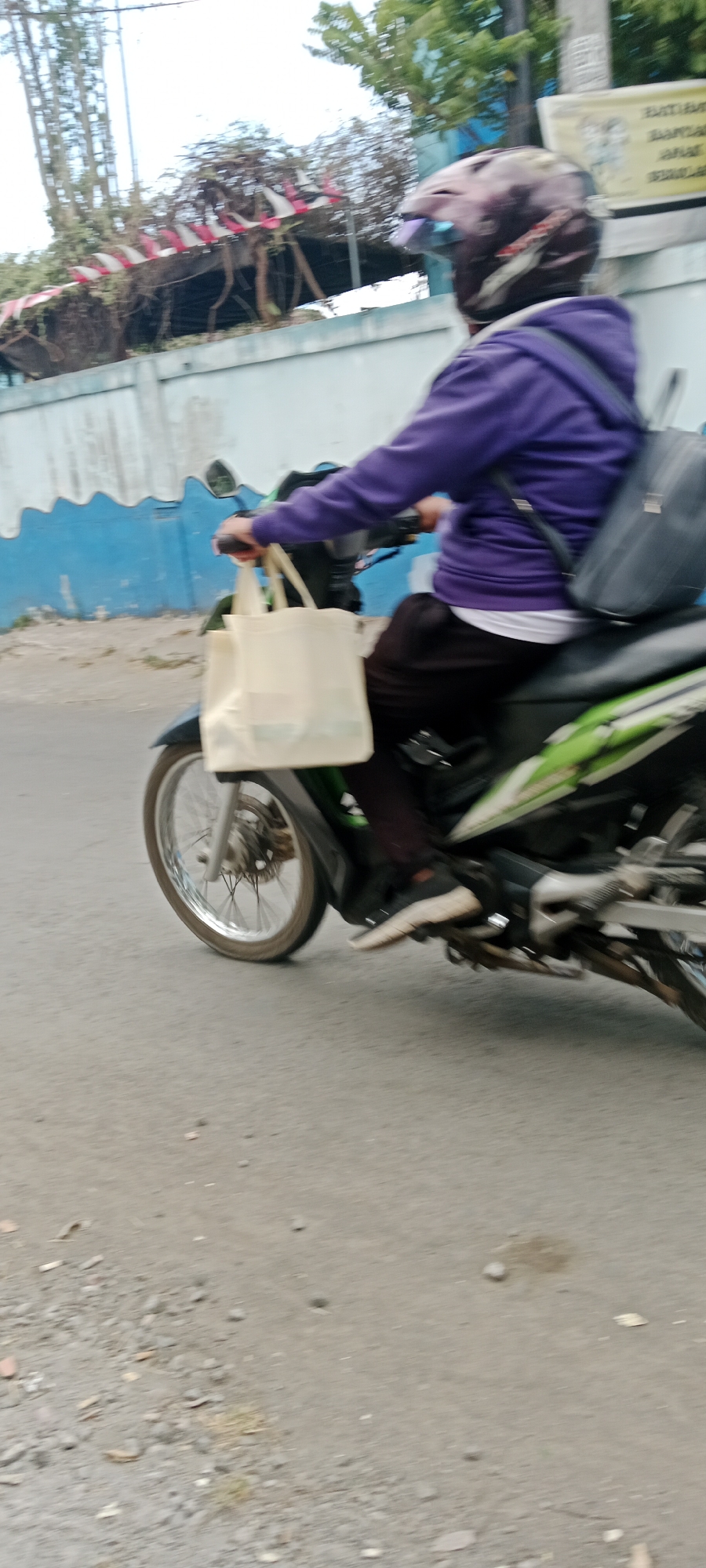 A person wearing a purple hoodie and a helmet is riding a motorcycle on a street in Purwomartani, Indonesia. The person is carrying a light-colored tote bag on their left arm and a black backpack on their back. The motorcycle has green and black accents. In the background, there is a light blue wall with a wavy pattern, some trees, and a white building with a sign. The ground is a paved road with some gravel and debris. The image is slightly blurred, indicating movement.