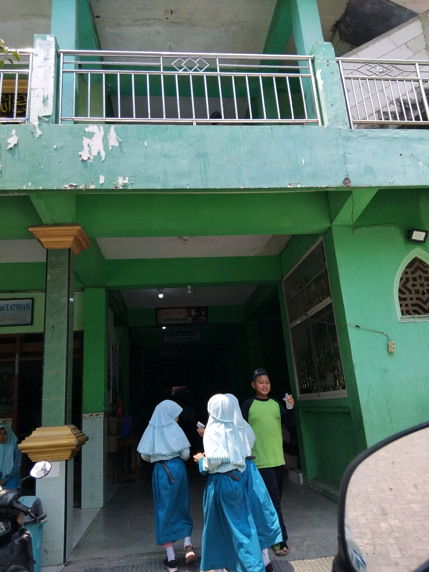The image depicts the exterior of a building, likely a school or religious institution, painted in a faded green color. The building has multiple levels, with a balcony on the upper floor featuring a metal railing. Peeling paint is visible on the exterior walls. In the foreground, three young individuals are present: two girls wearing light blue hijabs and blue dresses, and one boy in a green long-sleeved shirt and black pants, wearing a black cap. The boy is holding a water bottle. The girls appear to be walking away from the camera. To the left, a motorcycle is partially visible, along with a trash bin. Signs with text, some in Arabic script, are mounted on the walls. The scene is outdoors, with shadows indicating daylight. The architecture suggests a location in Southeast Asia, possibly Indonesia, given the clothing of the individuals. The overall impression is one of a functional, perhaps older, building in an urban or semi-urban setting.