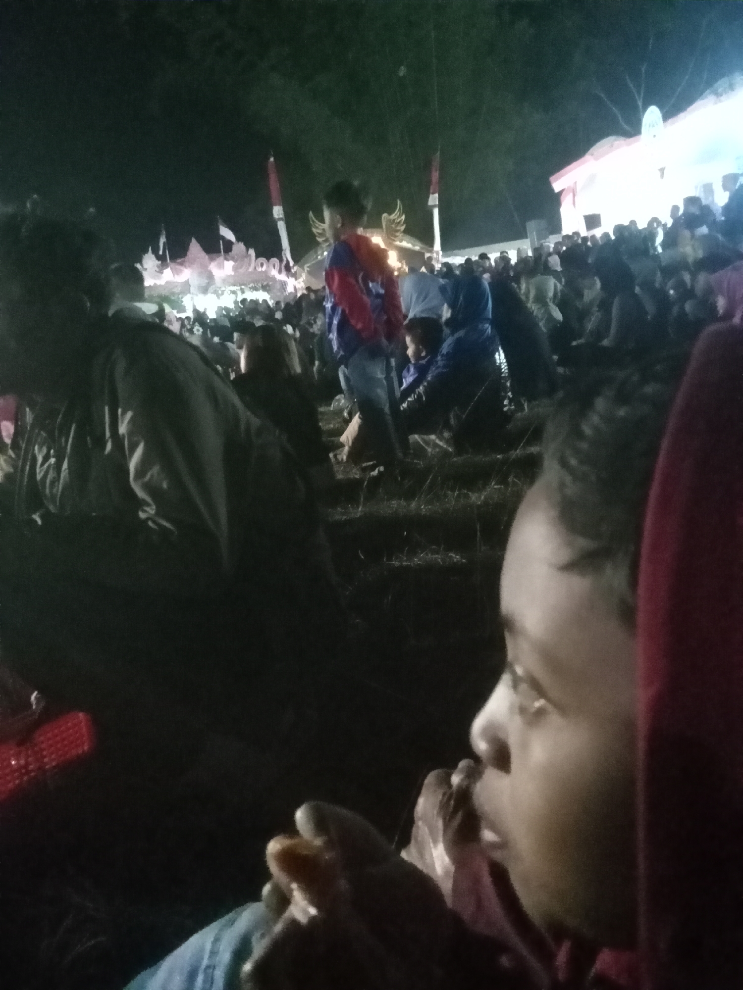 The image depicts a nighttime gathering of people, likely at an outdoor event in the city of Pule, Indonesia. The foreground is dominated by the profile of a young child, looking to the right and appearing to eat something. Their face is illuminated by an unseen light source. Behind the child, the scene opens up to a crowd of individuals, many of whom are silhouetted against bright lights. In the background, there are structures that appear to be stages or booths, decorated with what seem to be flags, including the Indonesian flag (red and white). The overall impression is of a festive or community event happening after dark. The environment appears to be an open area, possibly a field or public space, with some vegetation visible in the shadows. The lighting conditions suggest it is nighttime, with artificial lights providing illumination.
