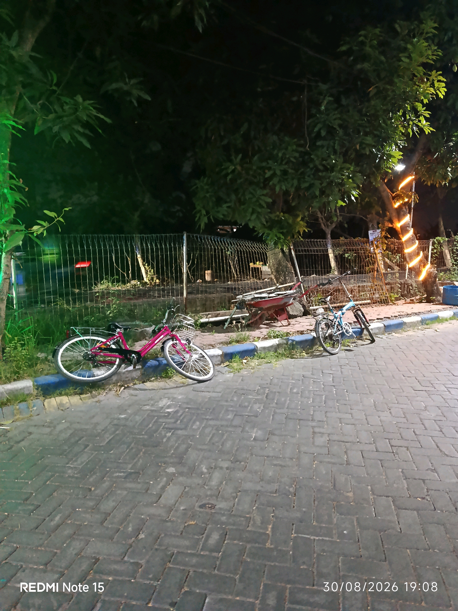 This is a nighttime outdoor photograph of a paved area with two bicycles. The bicycles are parked on a sidewalk with a blue and gray border. Behind the sidewalk is a metal fence, and beyond the fence are trees and some structures. One bicycle is pink and has a basket on the front and a rack on the back. The other bicycle is blue and appears to be a folding bicycle. A red wheelbarrow is visible behind the fence to the left. The trees are illuminated by strings of orange lights. The ground is paved with dark interlocking bricks. The bottom left corner of the image has the text "REDMI Note 15" and the bottom right corner has the text "30/08/2026 19:08". The location is identified as Keboansikep, Indonesia. There are no people visible in the image.