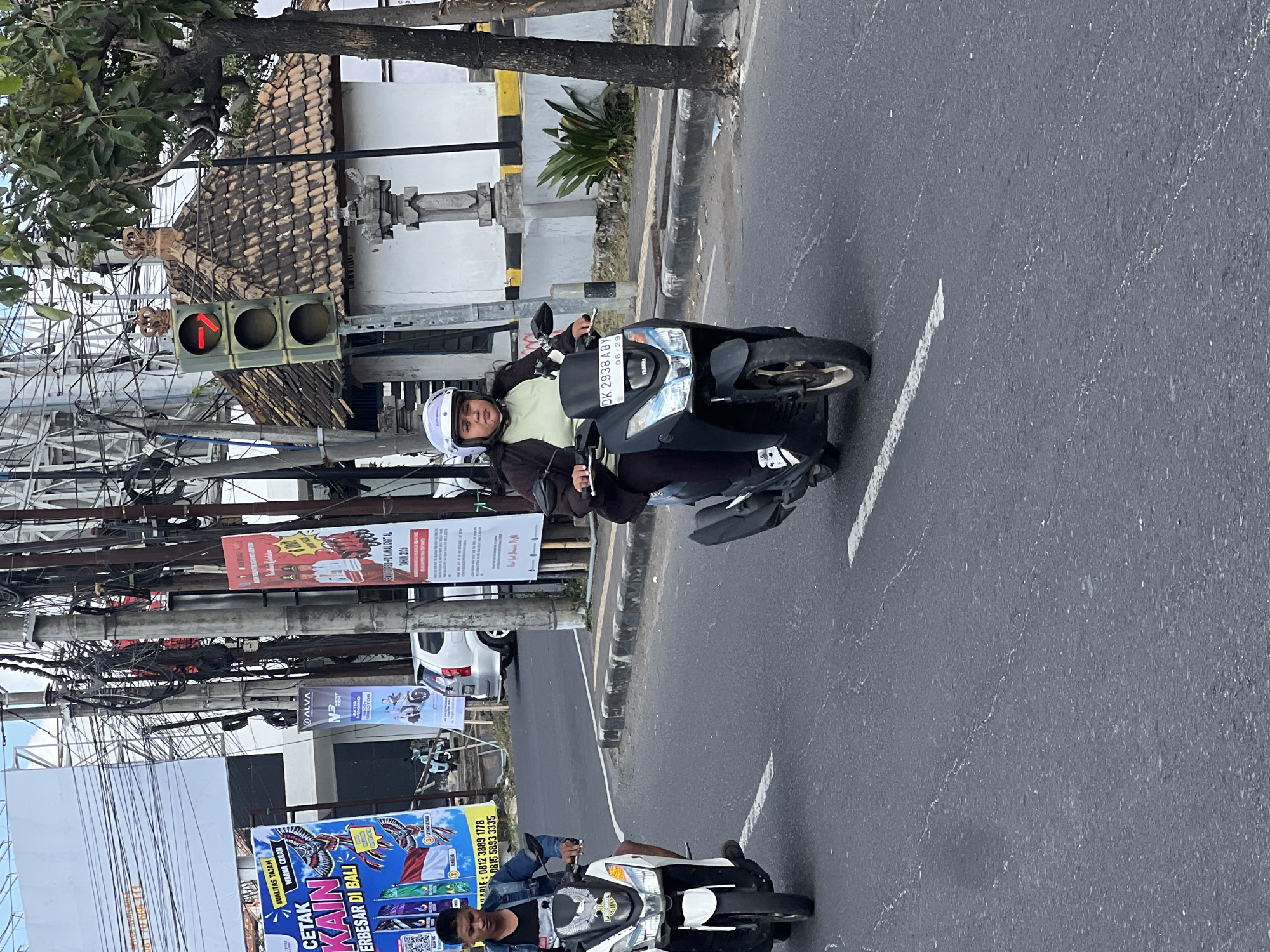 In Denpasar, Indonesia, two individuals are riding motorcycles down a paved road. The motorcycle on the left is white and is being ridden by a young man wearing a denim jacket over a dark t-shirt and shorts. The motorcycle on the right is black and is being ridden by a woman wearing a light green t-shirt, a dark jacket, dark pants, and a white helmet. The woman's motorcycle has a license plate that reads "DK 2938 ABY". A traffic light is visible in the background, showing a red arrow pointing to the right. There are utility poles with numerous wires and several signs in the background, indicating an urban environment. The road is marked with white lines and appears to be in good condition.