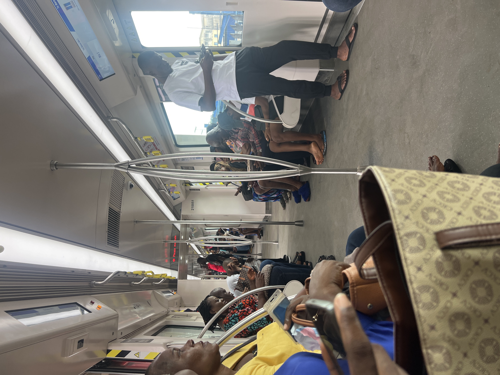 The image shows the interior of a modern train, likely the Lagos Rail Mass Transit system in Agbado, Nigeria. The train is populated with passengers, some seated and some standing, all appearing to be in transit. The train's interior is well-lit with overhead fluorescent lighting and features silver handrails and poles for passengers to hold onto.
In the foreground, a woman in a yellow top is partially visible on the left, with her hands holding a bag and a mobile phone. To her right, a woman in a colorful patterned top is seated, looking down at her phone. Several other passengers are seated in rows, their attire suggesting a casual environment. A man in a white t-shirt and black pants stands on the right side of the train, engrossed in his phone. Behind him, through the windows, urban buildings and infrastructure are visible, consistent with a city landscape.
The floor of the train is a light gray, speckled material, and the seats appear to be a light color. Digital screens are mounted above the windows, displaying train information. The overall impression is of a functional and active public transportation system in a Nigerian urban setting.