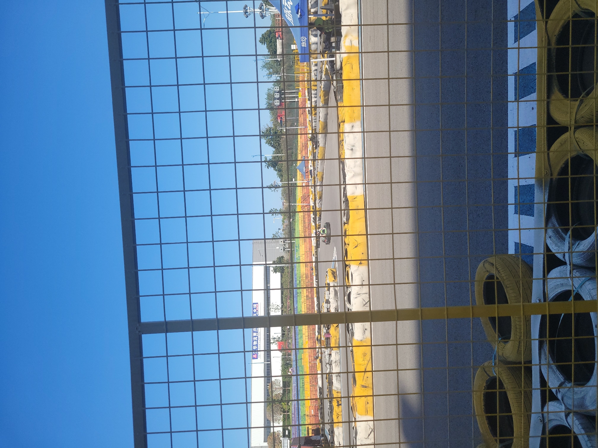 A go-kart track is visible in the city of Liangjiang, China, under a clear blue sky. The track features several go-karts in motion, with drivers wearing helmets. The track is bordered by yellow and white barriers, with colorful banners in the background. A large white building with Chinese writing is visible behind the track, suggesting it is a commercial or industrial area. A metal fence is in the foreground, partially obscuring the view of the track. Tires, some painted yellow, are stacked along the edge of the track, serving as additional protection. A blue canopy tent is set up trackside, with a person in a green uniform standing nearby. The overall scene depicts a recreational activity in an urban setting.