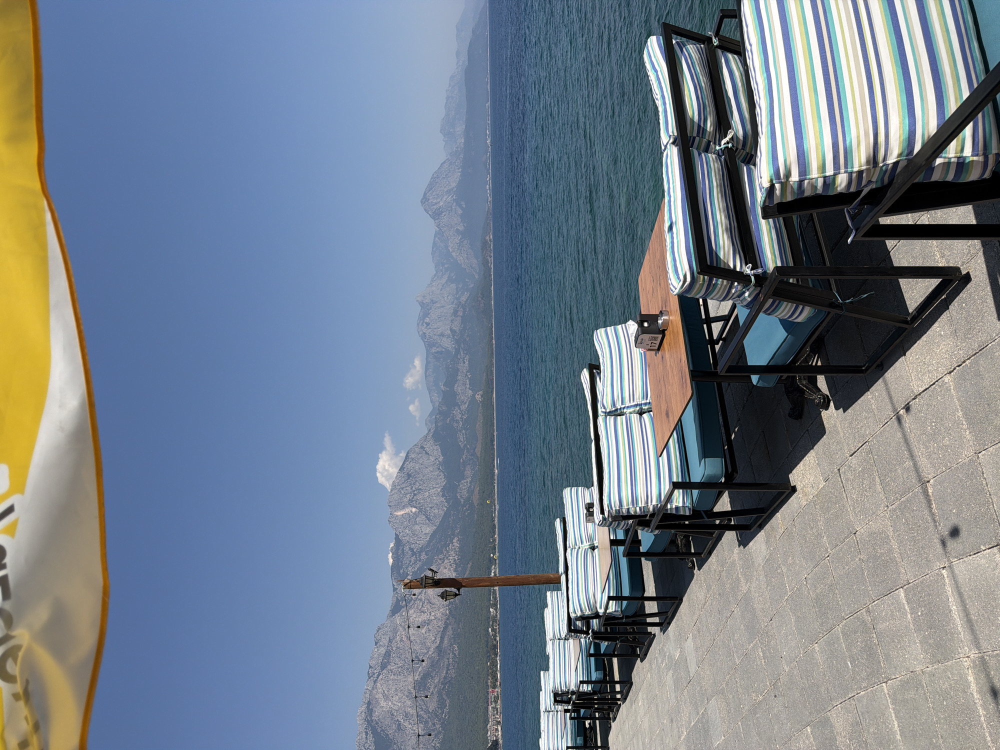 The image depicts an outdoor seating area of a restaurant or cafe situated on a paved walkway next to a body of water. The seating consists of black metal-framed benches with blue cushions and striped pillows featuring blue, green, and white patterns. Small wooden tables are placed between the benches. In the background, there is a calm, blue sea that stretches to the horizon, meeting a clear, bright blue sky. Beyond the water, a mountain range with rocky, rugged peaks dominates the landscape. The lower slopes of the mountains are covered in lush green vegetation, and a town with buildings is visible along the coastline. A wooden pole with a lantern and string lights is positioned to the left of the seating area. The overall scene conveys a serene and picturesque atmosphere.