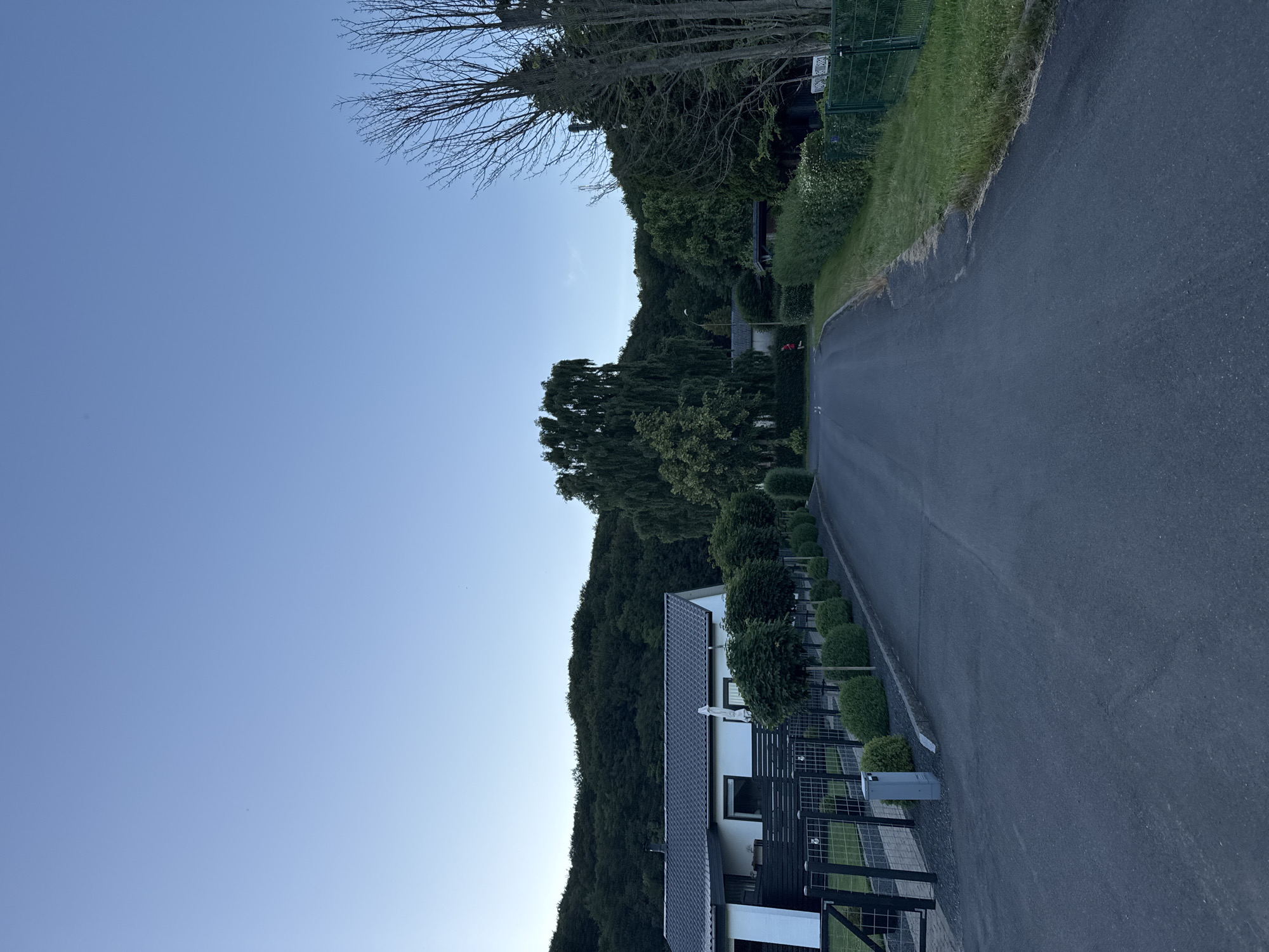 A paved road leads through a residential area with houses on the left and dense trees on the right, under a clear blue sky. A single person in red is jogging on the road, heading away from the camera. The house on the left is modern with white walls and a dark roof, bordered by a black fence and manicured hedges. The background features a steep, tree-covered hill. The scene suggests a suburban or semi-rural setting.