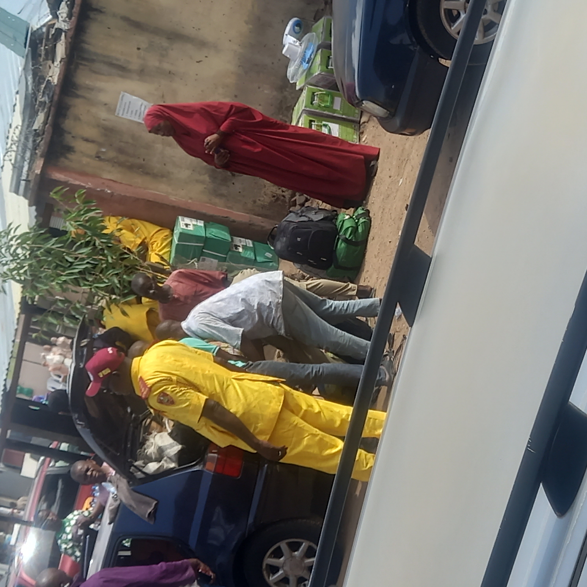 In Sardauna, Nigeria, the scene shows a group of people loading packages into the trunk of a dark blue car. There are at least six people visible: one woman in a red hijab standing nearby, and several men loading the packages. One man wears a yellow outfit and a red baseball cap, while another wears a light blue shirt. The packages are a mix of yellow and green. There are also fans in boxes next to the car and a black and green backpack. The setting appears to be an outdoor area next to a building.