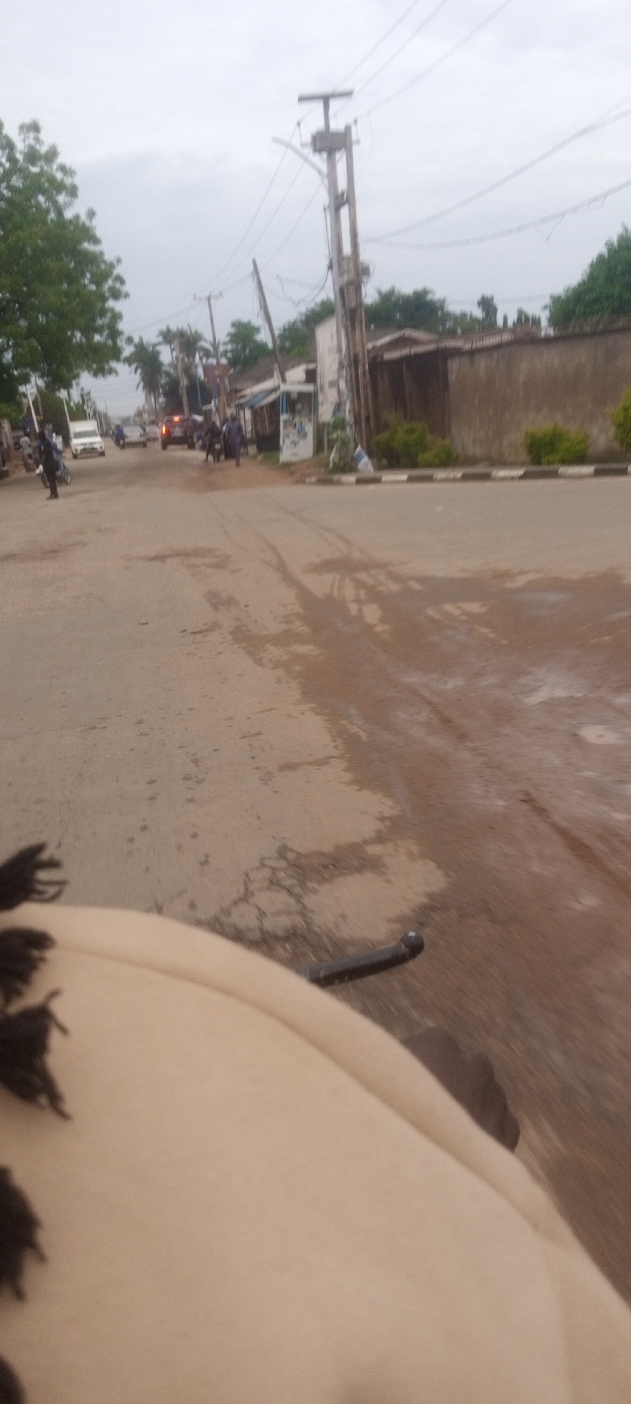 The image shows a street scene in what appears to be a residential area of Narayi, Nigeria. The road is paved but in poor condition, with visible cracks, patches, and areas that appear wet or damp. A white car, a red car, motorcycles, and pedestrians are present on the street. On either side of the road are buildings and walls, with vegetation present. Electrical poles with wires run along the roadside. The photo seems to have been taken from a moving vehicle, with part of the vehicle visible in the foreground.