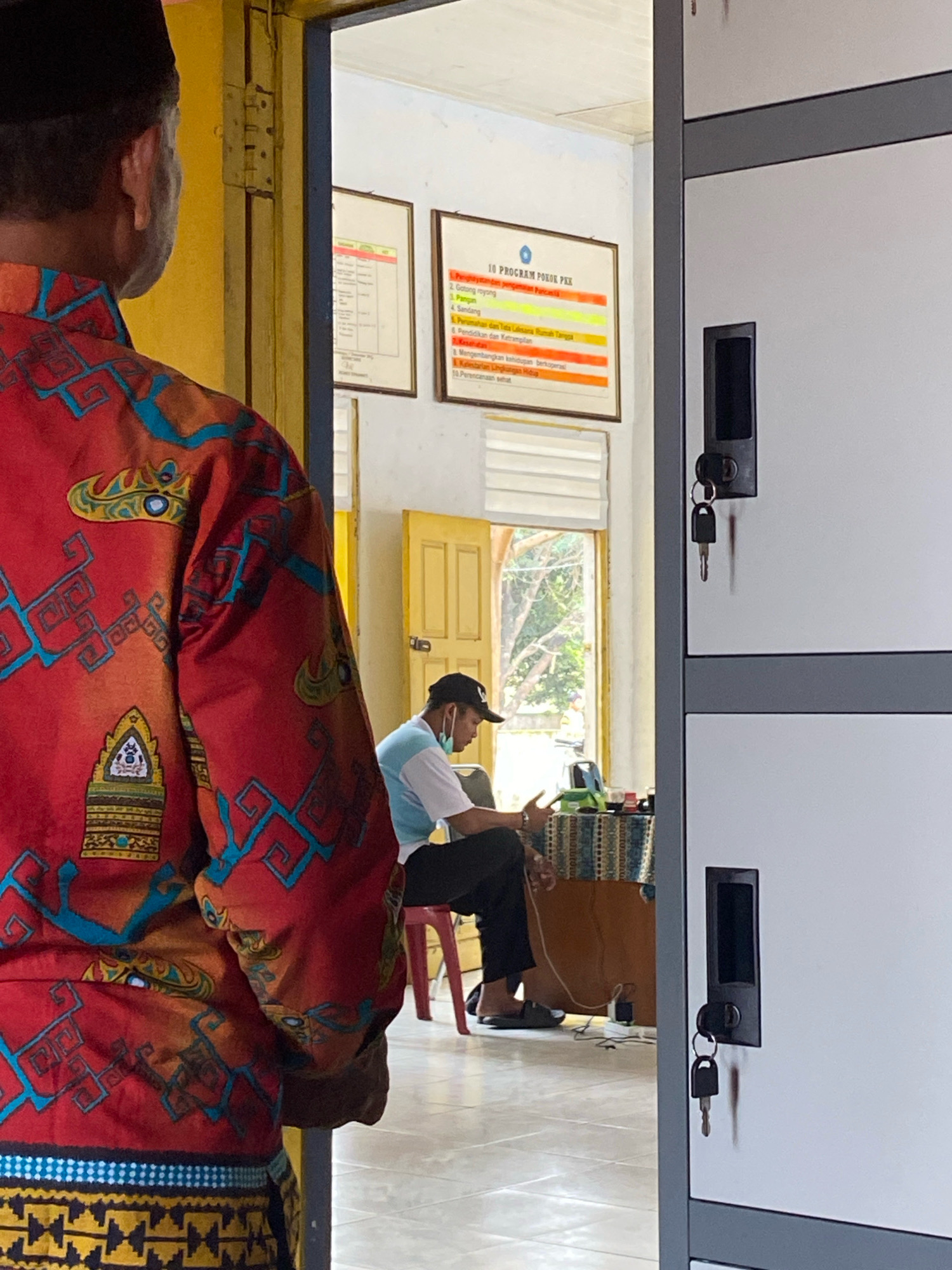 The image depicts an interior scene that appears to be an administrative office or a classroom in Mojo Pahit, Indonesia. In the foreground, a person is seen from the back, wearing a black peci hat and a red, ornately patterned shirt. The shirt features a repeating motif of blue geometric shapes and stylized, almost mythical creatures.
Further into the room, a second individual is seated at a desk, facing away from the camera. This person is wearing a black baseball cap, a light blue and white shirt, and dark trousers. They are also wearing a light-colored mask around their neck, suggesting a recent or pending mask use. The seated person is looking down at something on the desk and appears to be engaged in an activity, possibly administrative work or studying.
The environment includes a metal locker unit on the right side of the frame, with two visible compartments, each with a black handle and a key in the lock. Behind the seated person, there is a wooden desk with various items, including what appears to be a laptop or monitor, a woven mat, and some containers. A red plastic chair is visible next to the desk.
On the wall behind the desk, two framed documents are displayed. One of these, with the title "10 PROGRAM POKOK PKK", is clearly visible, featuring colored text in columns. A yellow door is open in the background, revealing a glimpse of the outside, where trees and sunlight are visible. The floor is tiled, and the walls are painted a light color, possibly white or cream. The overall impression is one of a functional, everyday space within a community or educational institution.