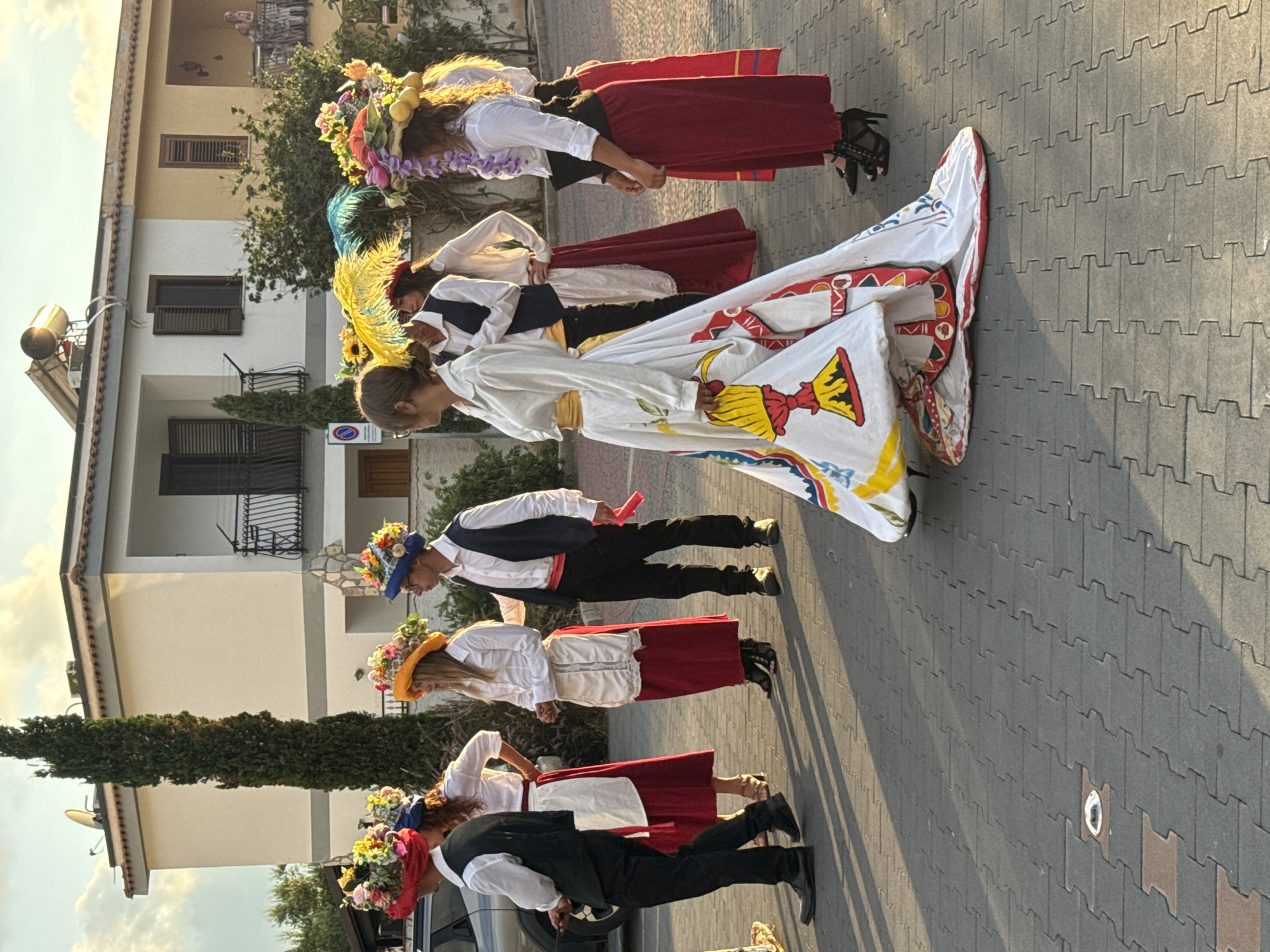 The image depicts a group of people in traditional costumes, possibly participating in a parade or festival, on a paved street in Isola delle Femmine, Italy. The architecture visible in the background suggests a residential area with buildings featuring balconies and awnings. The people are dressed in white shirts, with some wearing vests and red or white skirts. They are adorned with elaborate, flower-decorated hats. The person in the foreground on the right is wearing a white dress with a detailed, colorful pattern, featuring what appears to be a depiction of Mount Etna. The lighting suggests it is late afternoon or early evening, with long shadows cast on the street. The street is paved with interlocking bricks, and a visible drain cover is present in the foreground. A silver car is partially visible on the left side of the image. The overall atmosphere appears festive.