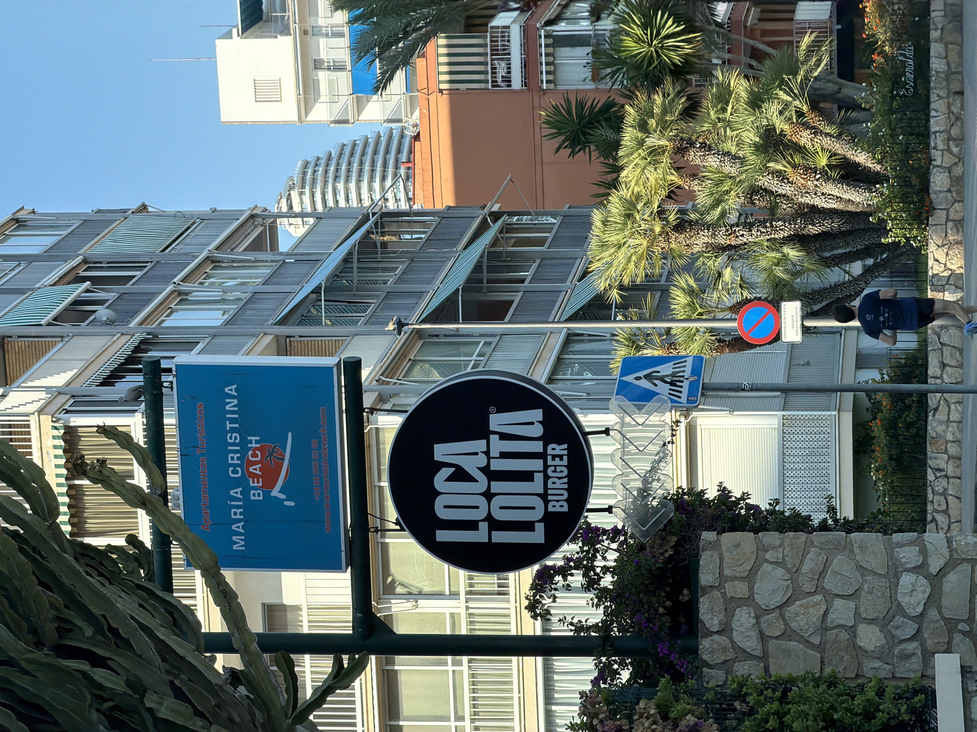 The image shows a street scene in Benidorm, Spain, with a focus on two signs mounted on a pole in the foreground. The upper sign, rectangular and blue, advertises "Apartamentos Turisticos MARIA CRISTINA BEACH," featuring a stylized image of a beach umbrella and a palm tree. Below this, a circular black sign reads "LOCA LOLITA BURGER" in white text.
Behind the signs, an apartment building with multiple balconies and green striped awnings is visible. To the right, palm trees and other greenery populate the scene. A pedestrian crossing sign with a blue background and a white silhouette of a person walking is also present. Further down the street, a person is seen jogging away from the camera. The sky is clear and blue. The overall environment suggests a coastal urban setting in a warm climate.