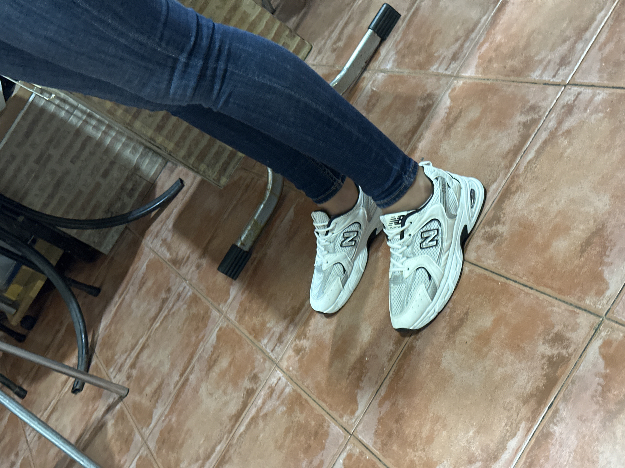 A person wearing blue jeans and white New Balance sneakers stands on a tiled floor. The sneakers have the characteristic "N" logo on the side. The floor is made of terracotta-colored square tiles with visible grout lines. In the background, metal chair legs are visible, along with part of a striped wall and some other furniture. The scene appears to be indoors, possibly in a home or a commercial space. There is no indication of the specific location in Alicante/Alacant, Spain.
