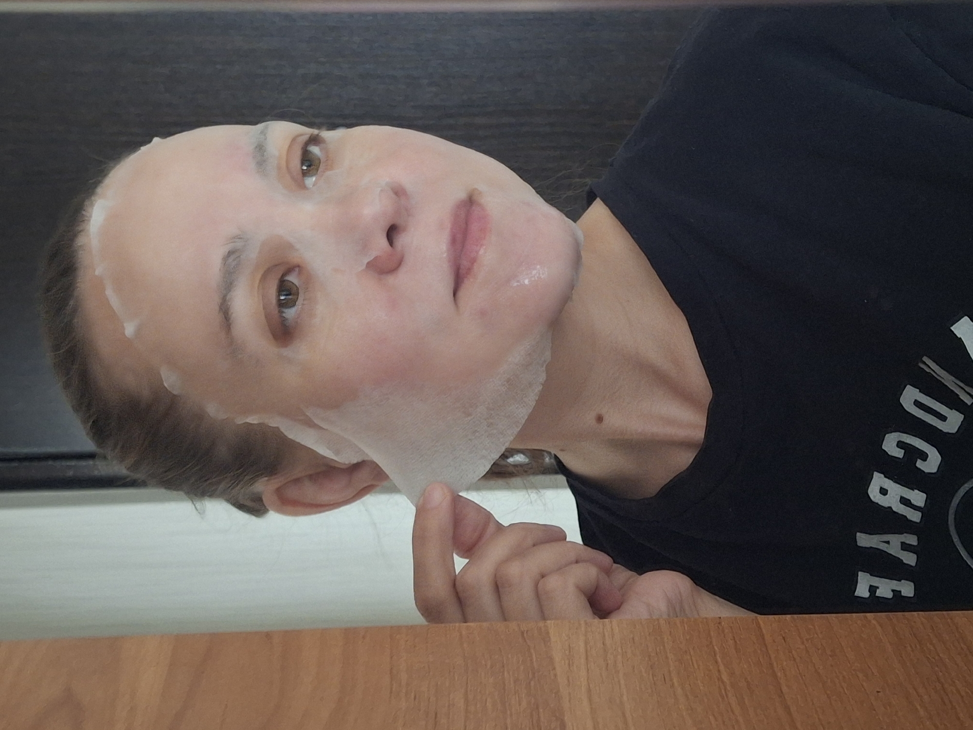 A woman is lying on her back with a sheet face mask on. She is wearing a dark T-shirt. Her hand is near her face, and she is looking directly at the camera. The background includes a dark surface above and a light wood surface below. Based on the location provided, this image was likely taken in Chaikovsky, Russia.