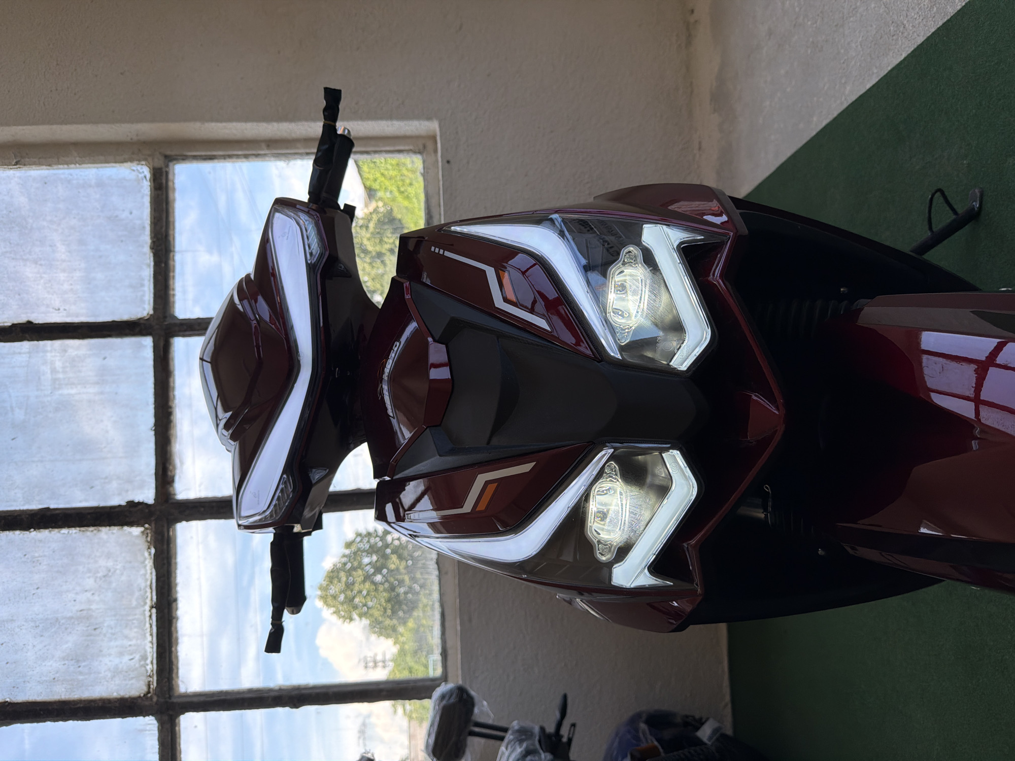 A red scooter is parked on a green carpet in front of a large, dirty window. The scooter's headlights are on and illuminating the area. The window looks out onto trees and a cloudy sky. The scene appears to be indoors, possibly in a garage or showroom. There are no people in the image.