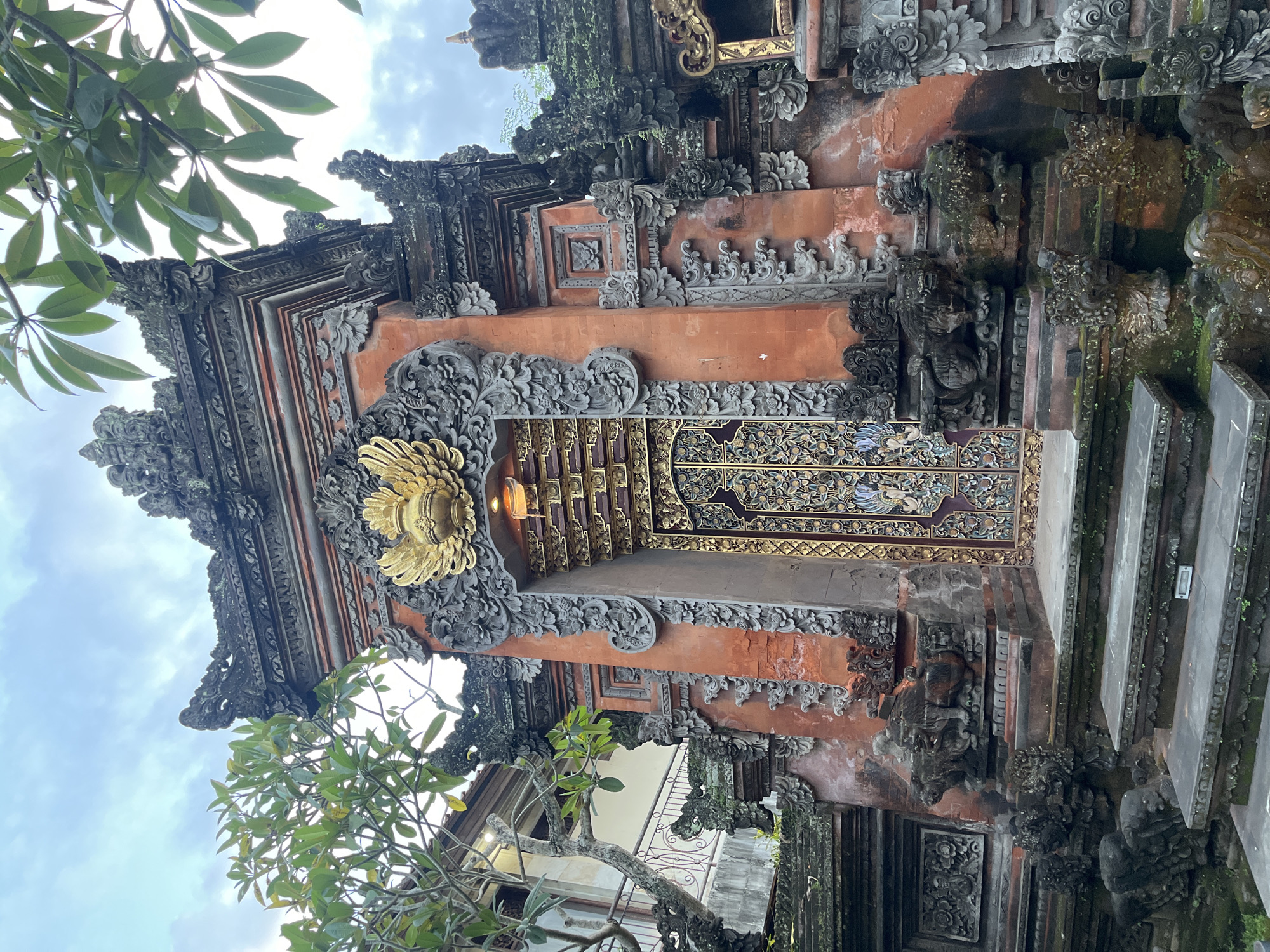 The image shows a richly ornamented Balinese temple gate located in the Ubud District of Indonesia. The gate is predominantly constructed from brick and stone, exhibiting a patina of age. Complex carvings adorn the structure, including floral motifs, geometrical patterns, and figures of deities or guardians. A large, gilded floral emblem centers the top of the entrance. A pair of elaborately decorated doors can be seen inside. The surrounding area appears to be lush with tropical vegetation, including tree foliage that partially obscures the upper portion of the gate. Steps are on the right side, leading to the top of the structure. The sky is overcast.