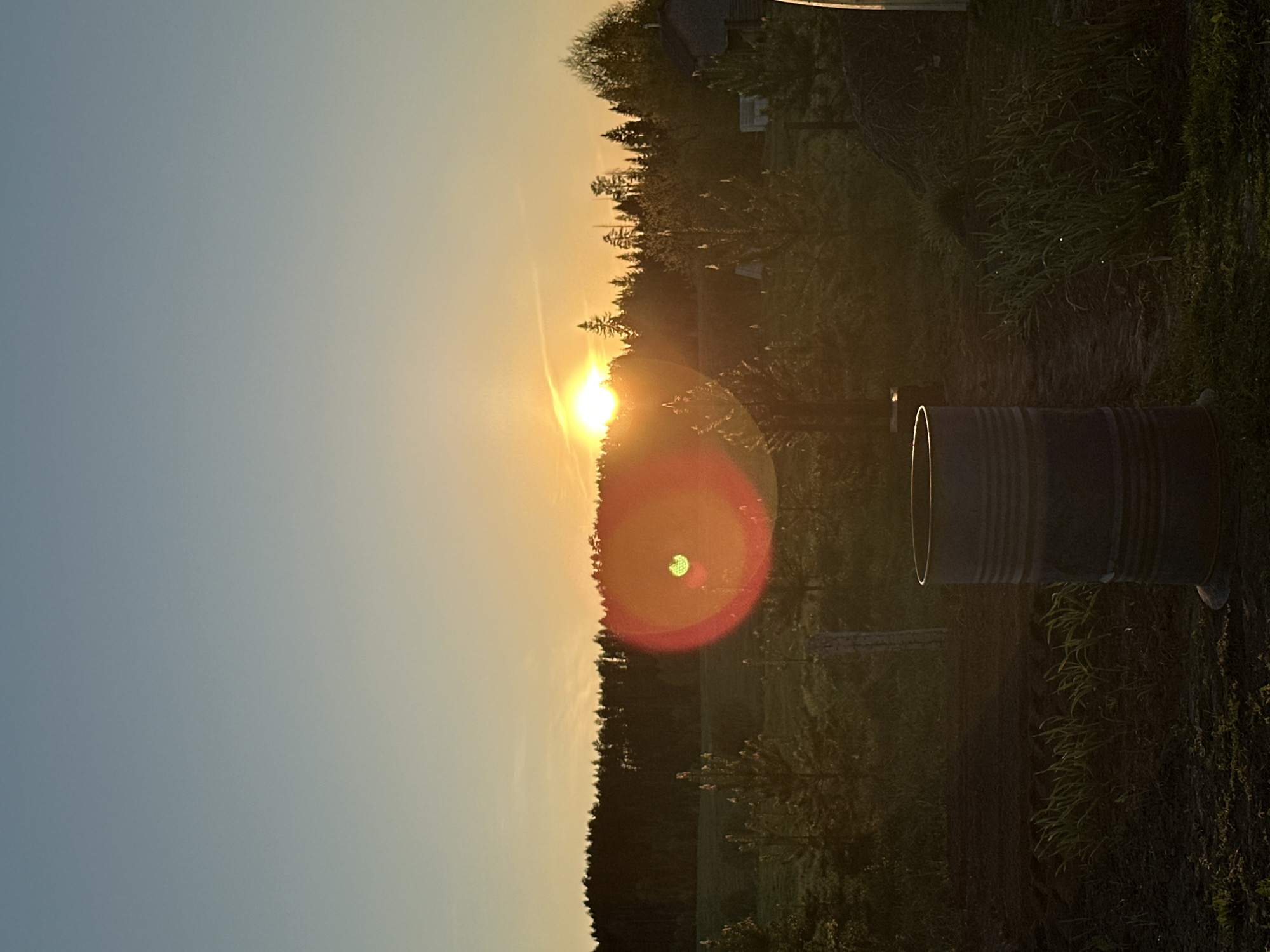 The image shows a sunset in Среднее Кечево, Russia. A cylindrical metal barrel is in the foreground, resting on the ground amidst grass and foliage. Behind the barrel, the landscape transitions into a field and a line of trees silhouetted against the setting sun. The sun is bright, creating a lens flare in the image. The sky shifts from a light blue to an orange hue near the horizon. The overall scene is rural, with a mix of natural elements and a man-made object.