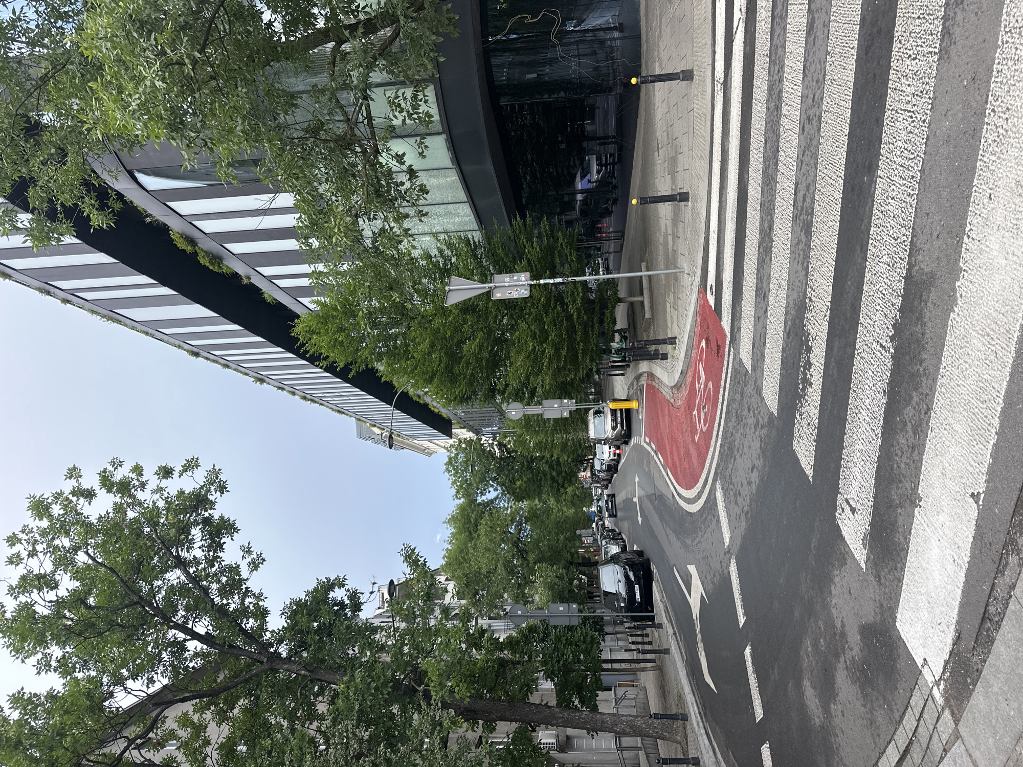 The image depicts a city street scene in Warsaw, Poland. In the foreground, a pedestrian crosswalk with white stripes spans across the asphalt road. To the left of the crosswalk, a red-painted lane, likely designated for bicycles, curves into the street. A black SUV is parked along the left side of the road, and a white car is visible further down the street. Tall trees with lush green foliage line the left side of the street, casting shadows on the pavement.
Dominating the right side of the image is a modern building with a dark facade and vertical white accents. Its reflective windows show glimpses of trees and the sky. The building has multiple stories, and balconies with greenery are visible on upper levels. A sidewalk runs along the front of this building, bordered by black bollards topped with yellow spheres.
The sky is overcast with a pale blue hue. The overall impression is that of a contemporary urban environment with a blend of modern architecture and mature trees. The street appears to be clean and well-maintained. There are no people visible in the frame.