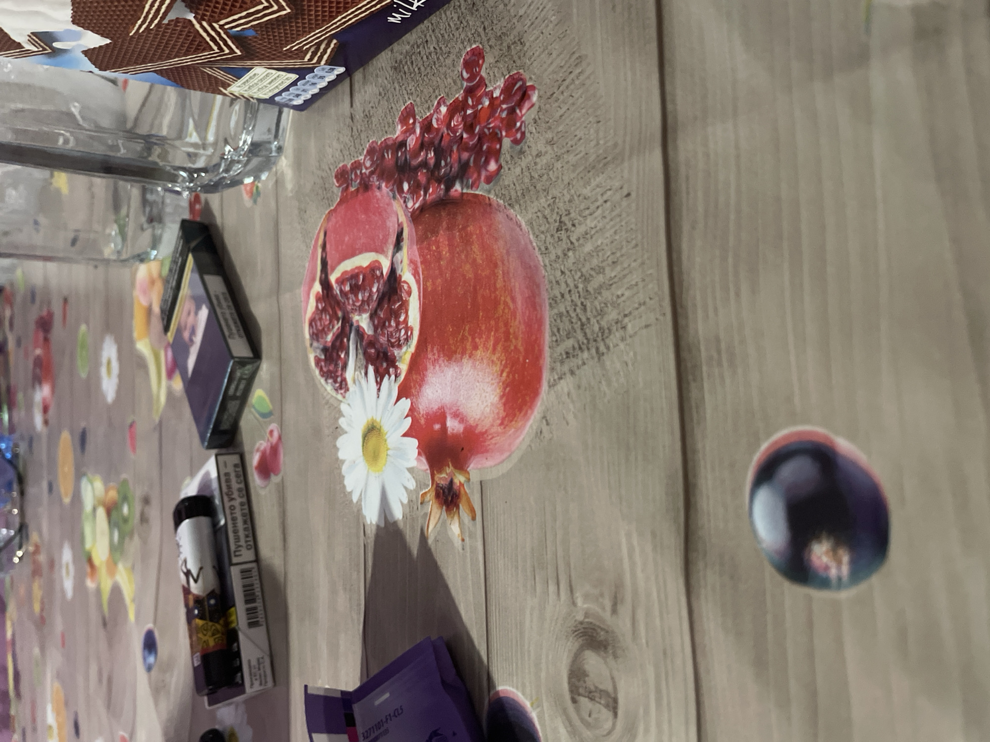 The scene is an indoor tabletop setting in the city of Ihtiman, Bulgaria. The table surface features a light brown, wood-grain patterned overlay, under which a colorful, abstract floral or circular pattern is visible, suggesting a tablecloth.
In the foreground, a large, multi-component paper cutout of a pomegranate is centrally placed. This cutout depicts a whole red pomegranate, another partially sliced open revealing red arils, and a scattered pile of individual arils extending to the right. A white daisy flower cutout with a yellow center rests on top of the whole pomegranate.
To the upper left of the pomegranate, a dark-colored disposable vape pen with gold and black designs and a white tip bearing a "W" logo is positioned atop a white cigarette pack. The cigarette pack displays a barcode and Bulgarian text which translates to "Smoking kills - quit now." Further left and slightly forward, a smaller cutout of a dark purple, berry-like fruit is visible on the table surface, exhibiting a slight red edge.
In the upper right quadrant, a clear glass drinking vessel with vertical ridges is partially visible behind the pomegranate cutout. To its right, a purple and brown packaged snack item, displaying layered wafer patterns and "milk" in white script, is also partially shown. In the lower-left corner, a crumpled purple packaging element with white text "3271101-FI-C1S" is present.
No individuals are present in the image, and no specific actions are underway beyond the static arrangement of these items.