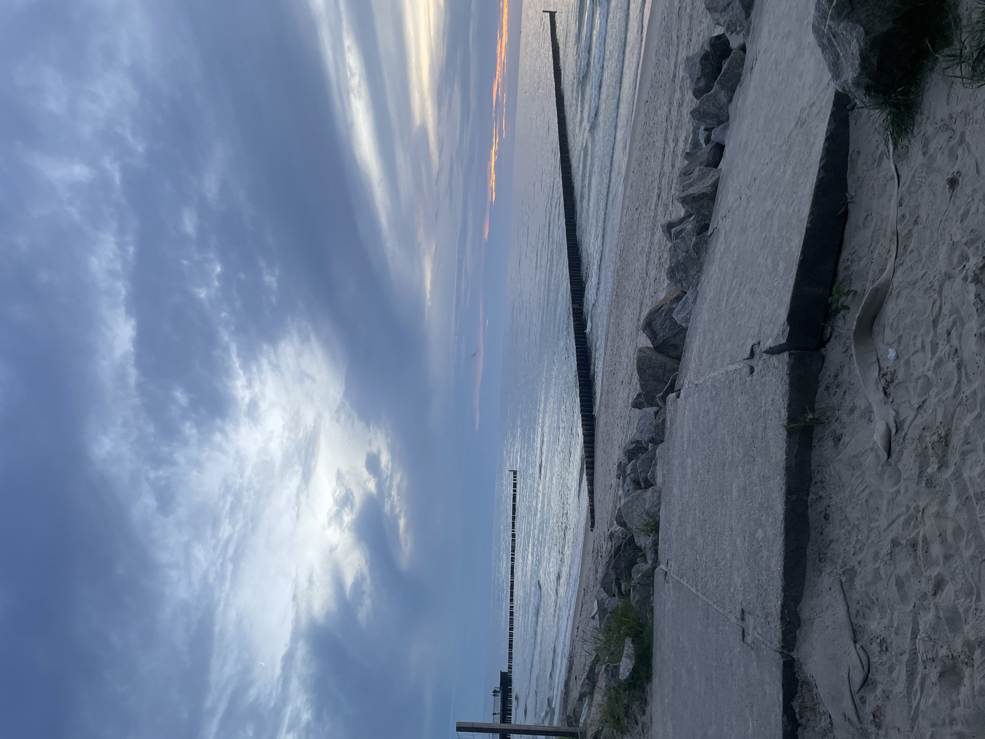 This is a wide shot of a beach in Kołobrzeg, Poland, at dusk. The sky is filled with dramatic, layered clouds in shades of grey and blue, with hints of orange and pink on the horizon where the sun is setting. The sea is calm, with gentle waves lapping at the sandy shore. Several rows of wooden groynes, likely breakwaters, extend out into the water, creating a rhythmic pattern against the sea. In the foreground, a concrete pathway lies partially on the sand, with rocks and some sparse vegetation scattered around it. There are no visible people in the immediate foreground or on the beach. A distant pier-like structure with figures on it can be seen to the left. The overall atmosphere is serene and slightly melancholic, characteristic of a fading day.
