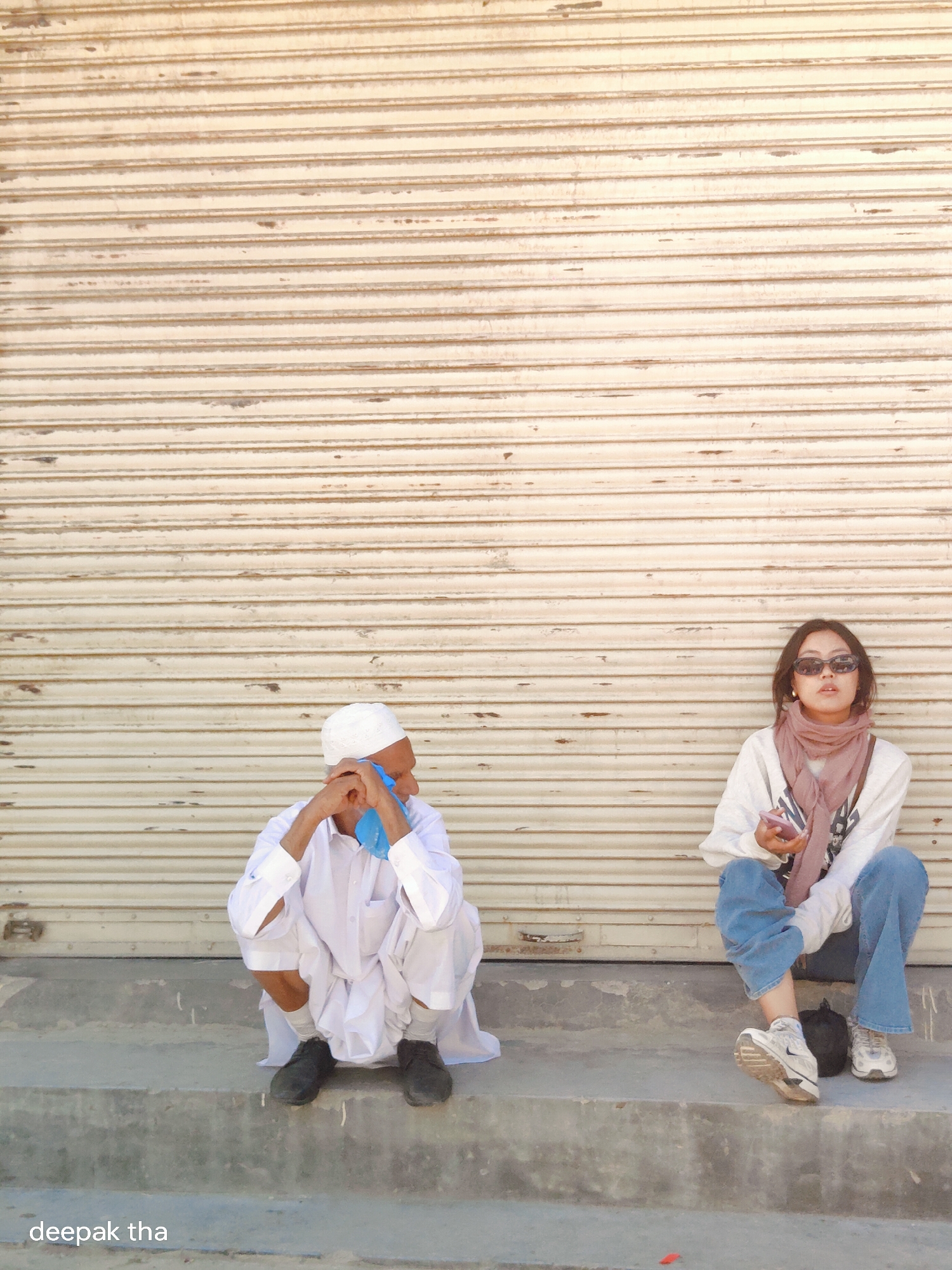 In Leh, India, two individuals are seated on concrete steps in front of a closed, corrugated metal shuttered storefront. On the left, a man wearing a white thobe and skullcap is crouched, holding a blue cloth to his face. To his right, a young woman with sunglasses and a pink scarf is seated, looking towards the viewer and holding a smartphone. The environment appears to be an outdoor urban setting, likely a street or alleyway.
