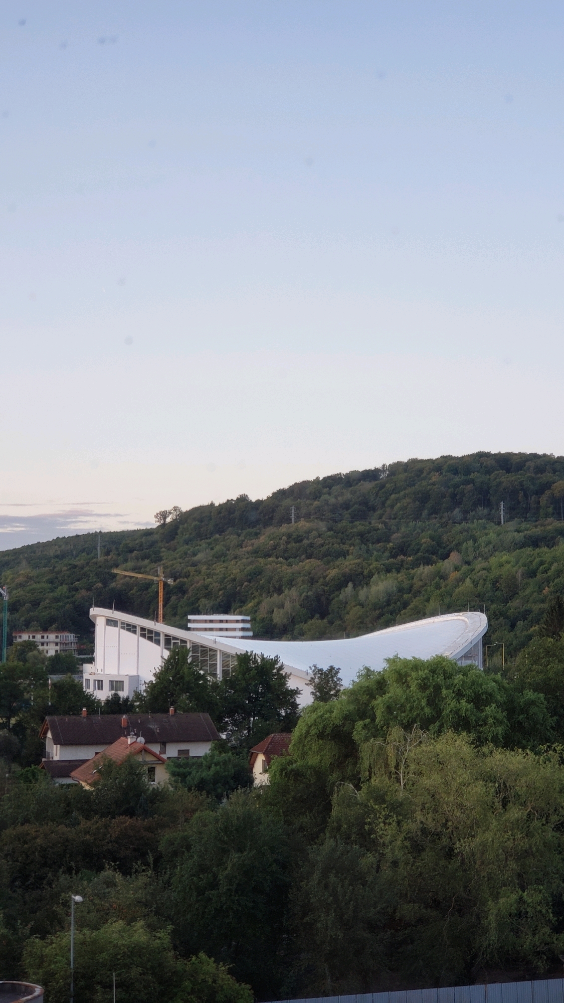 This is a vertical, eye-level, outdoor shot taken during daylight, showing a panoramic view of a modern building complex with a curved white roof, located on the side of a lush, tree-covered hill in Prešov, Slovakia. The building is white with large windows and a prominent white, wave-like roof structure. To the left of the main building, there is a construction crane. In the foreground, several houses with red-tiled roofs are nestled among dense green trees. The sky is a pale, cloudless blue. The overall scene depicts a blend of urban development and natural landscape.