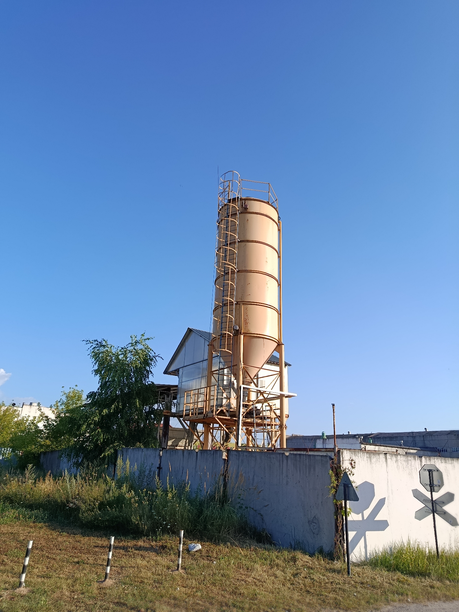 A tall, cylindrical, tan silo with a ladder on its side stands prominently against a clear blue sky. It is part of an industrial facility in Svietlahorsk, Belarus, featuring a sloped-roof building behind it and a network of metal supports and platforms. In the foreground, a rough concrete wall runs horizontally across the frame, partially covered by overgrown grass and weeds. Three black and white striped posts are visible in the grass in front of the wall. To the right of the wall, a white surface displays shadows of traffic signs, including a pentagon shape and a cross shape, suggesting proximity to a road or railway crossing. Additional industrial buildings are visible in the background.