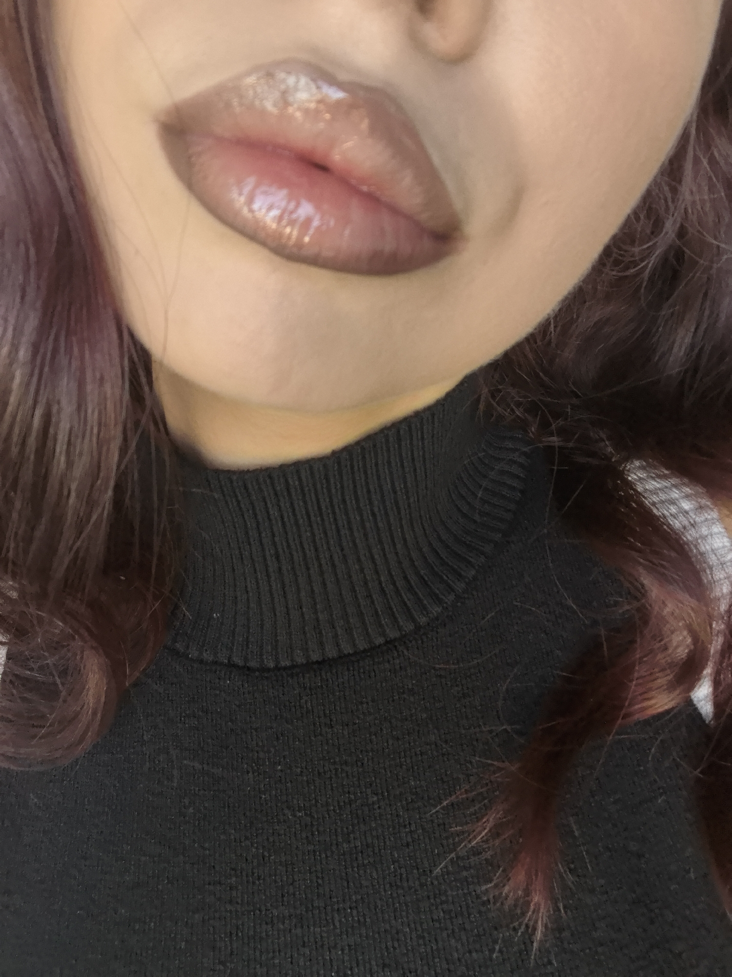 A close-up shot depicts a person wearing a black turtleneck sweater. The focus is on their lips, which are adorned with a glossy, taupe-colored lip product. The person has hair with burgundy tones, which is visible on both sides of the frame. The setting or location is not discernible from the image.