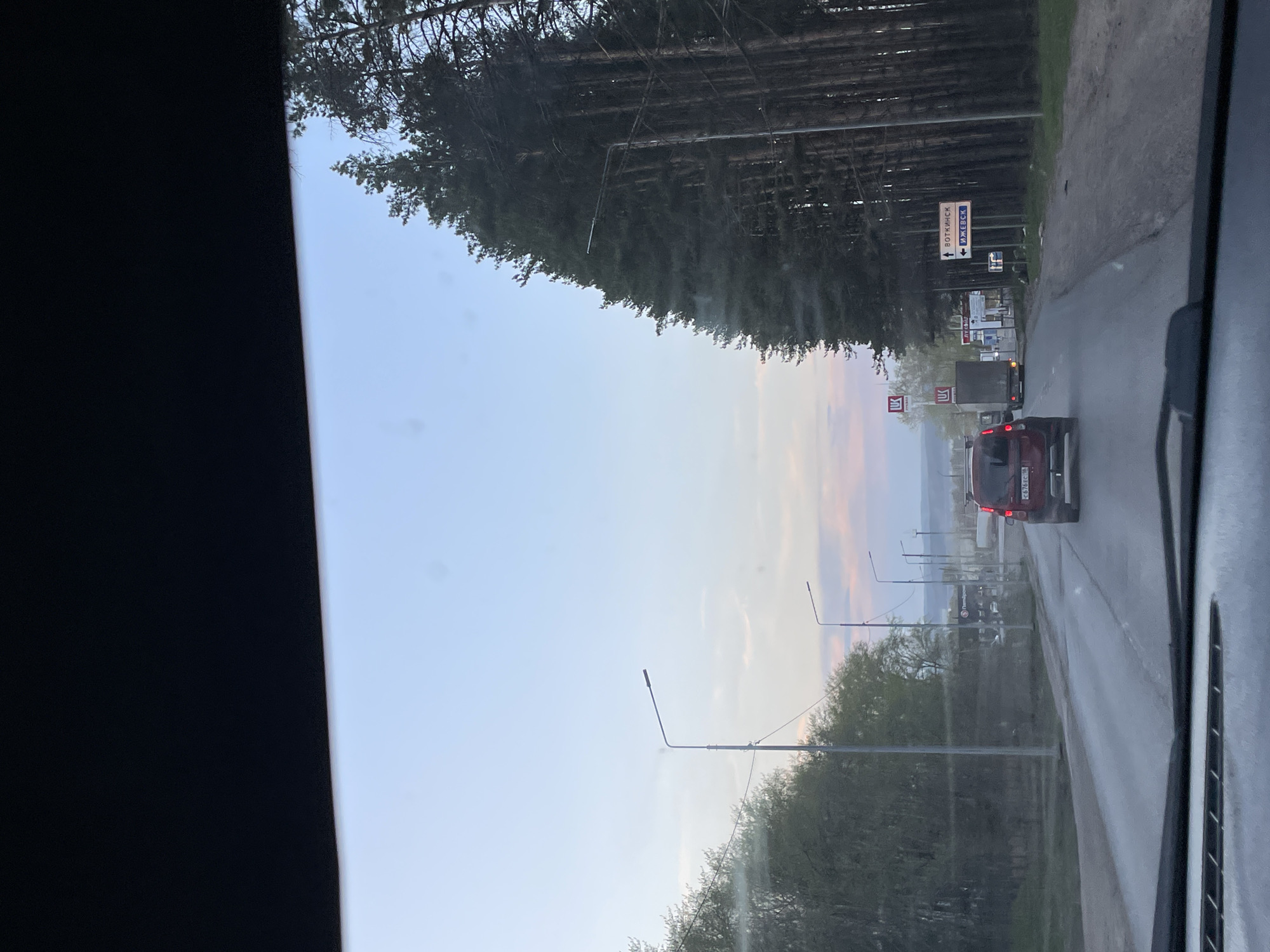 The image shows a view from inside a vehicle, looking out onto a road. In the distance, there is a red car and a truck. Road signs are visible near the side of the road. There are trees alongside the road, with a mix of dense forest and smaller trees. Streetlights line the road. The sky is visible, with colors suggesting either dawn or dusk. The location is likely a road on the outskirts of a city, with signs pointing towards Votkinsk and Izhevsk.