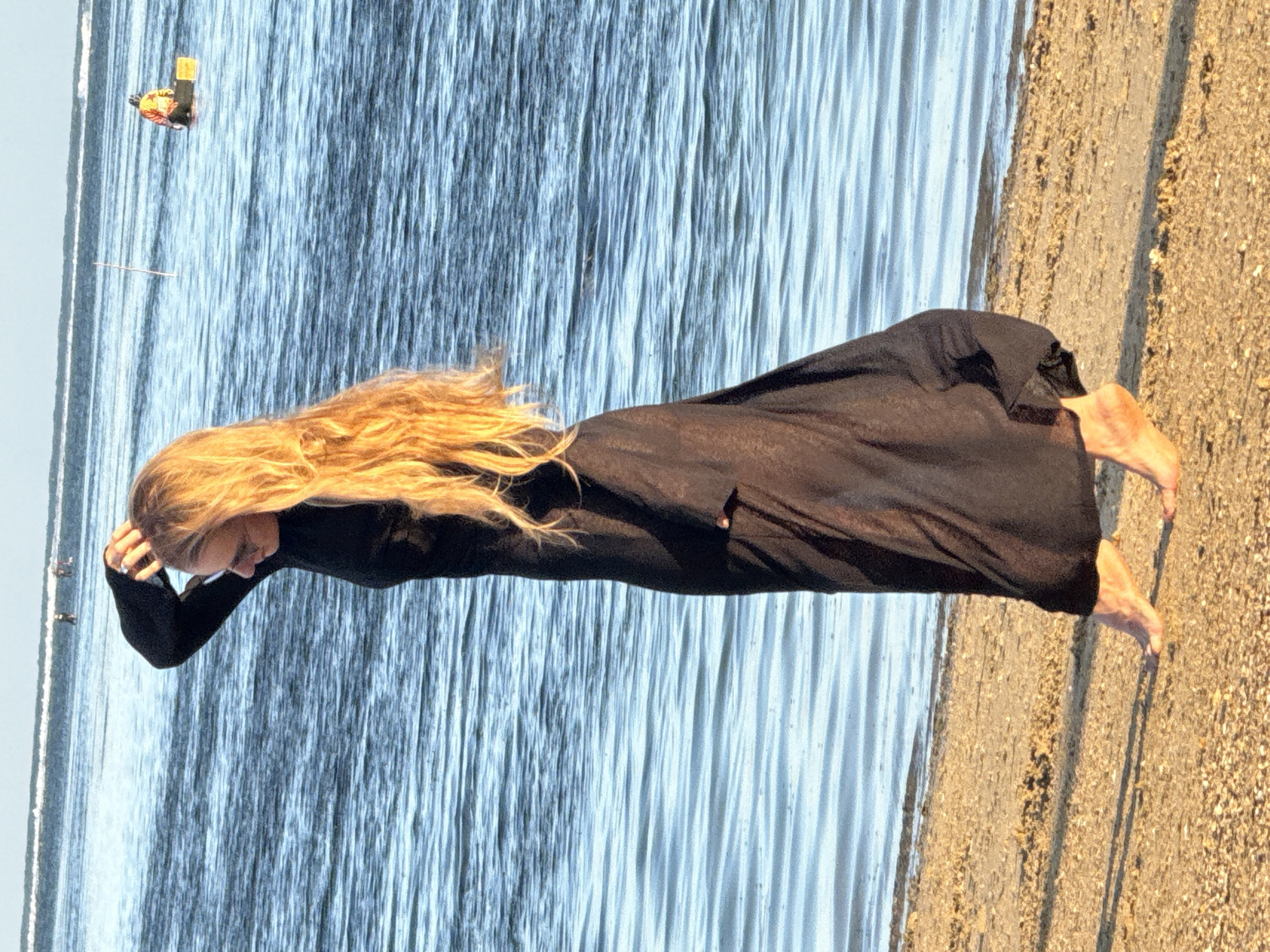 A woman with long blonde hair is standing on a sandy beach, her body angled to the side and her hand touching her head. She is wearing a long black dress. The ocean is behind her, with calm blue water. In the distance, there are several people in the water and a person in a yellow vest on a small raft. The sun is casting a warm glow on the sand and water. This scene is not located in Denpasar, Indonesia.