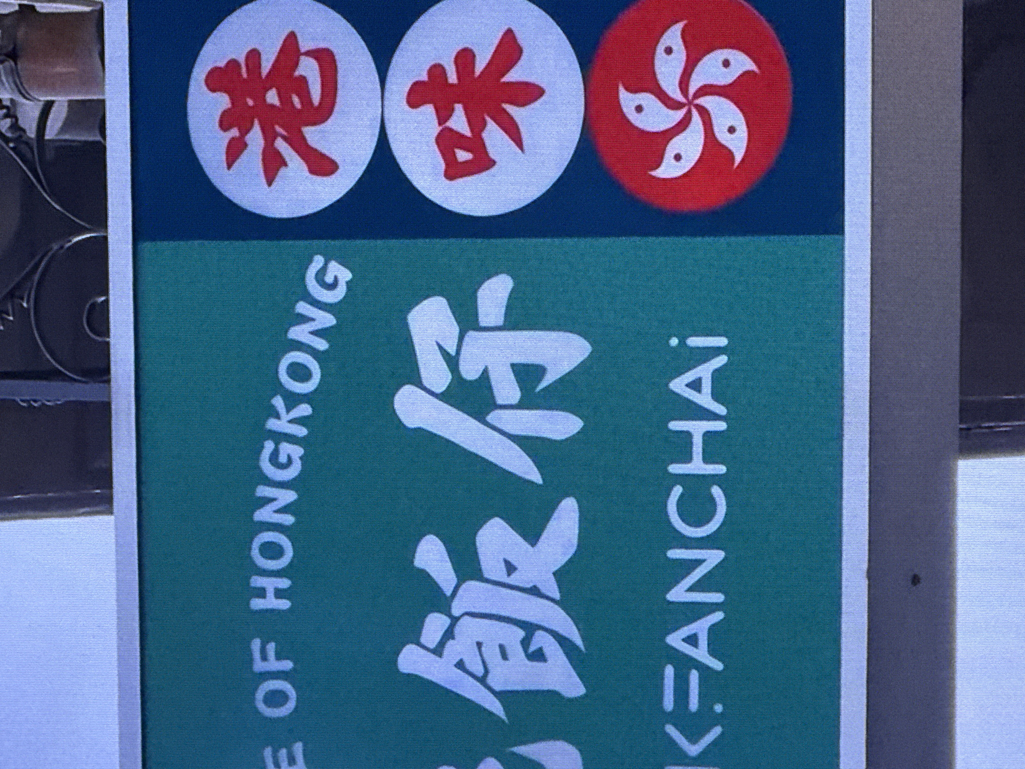 A sign with text and symbols is visible. The sign has a green background on the left and a dark blue background on the right. White text on the green background reads "E OF HONGKONG" and partially visible characters that appear to be Chinese. Below this, white text reads "K=FANCHAR". On the dark blue background, there are three circular elements. The top circle contains a red Chinese character. The middle circle also contains a red Chinese character. The bottom circle contains a red and white symbol that resembles a flower or a star, which is the Bauhinia blakeana, the floral emblem of Hong Kong. The sign is mounted outdoors, with a blurred background that suggests an urban environment. The image is taken from a slightly elevated angle.
