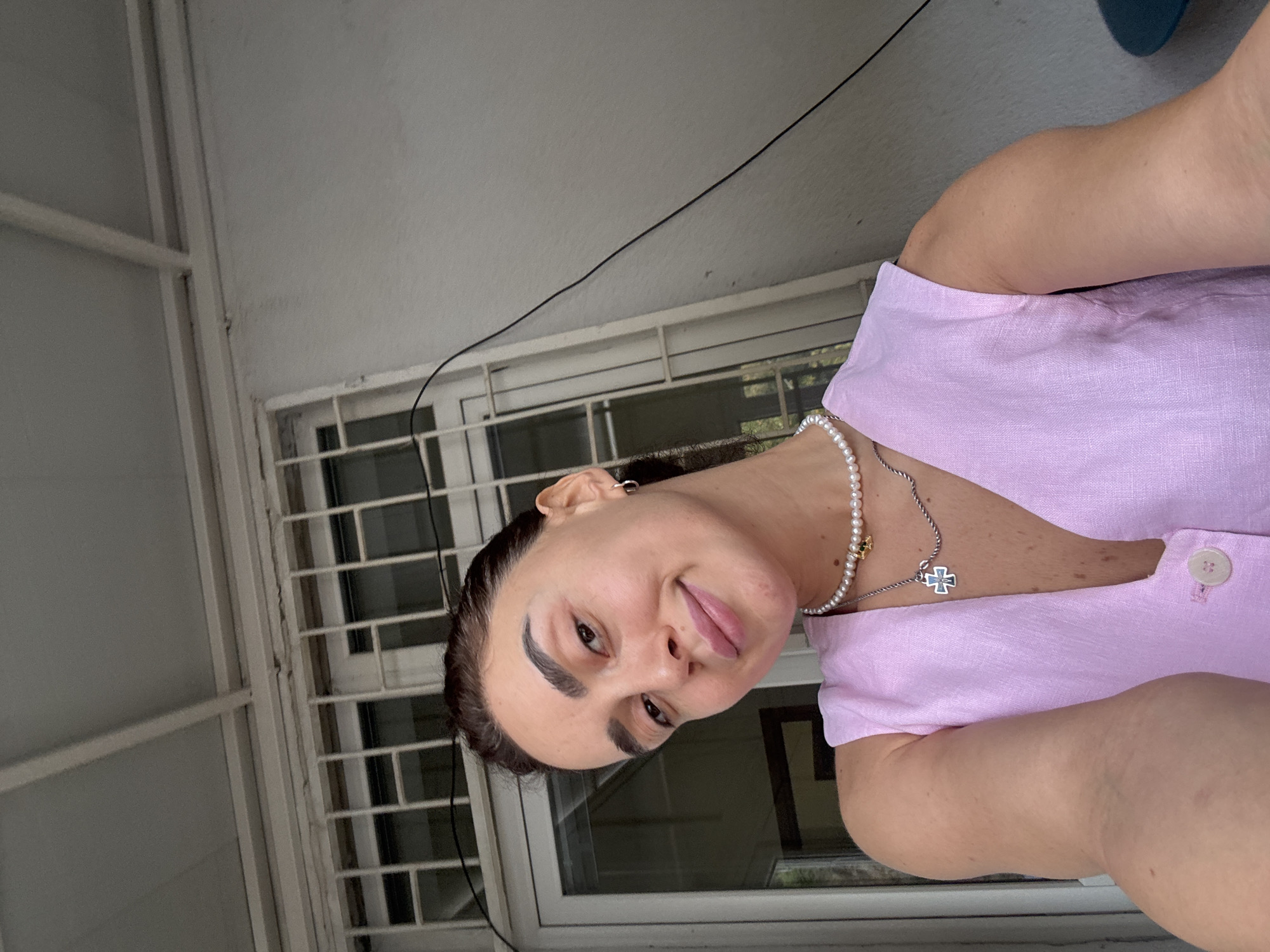 A young woman with dark hair pulled back is taking a selfie outdoors. She is wearing a light pink, sleeveless V-neck top with two buttons at the bottom. She has on a pearl necklace with a silver chain and a cross pendant. Her left arm is extended, holding the camera. The background shows a white-painted wall and a window with white bars. The environment appears to be a residential area, potentially in Tbilisi, Georgia, given the architectural style of the building and window grating.